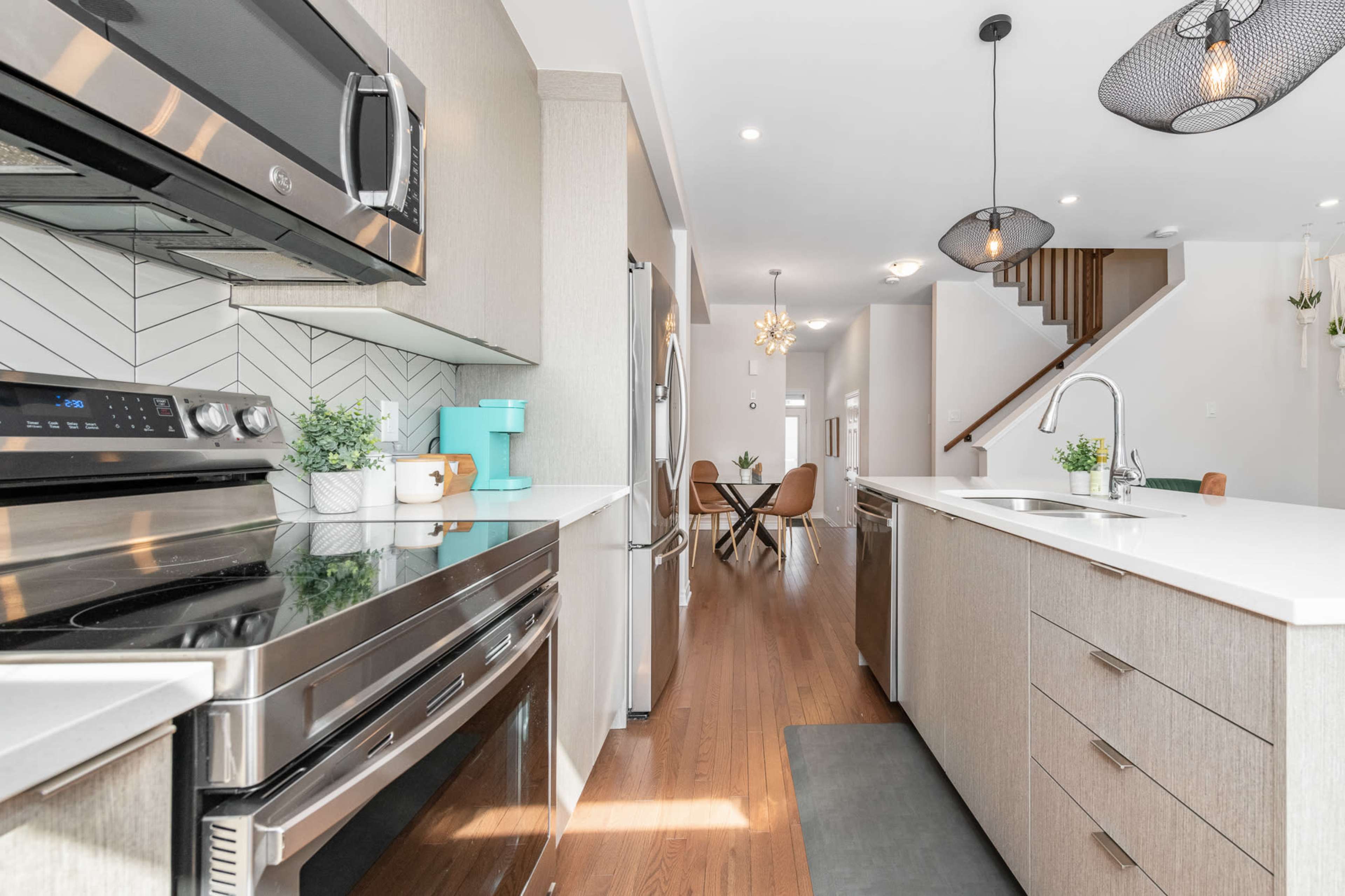 A modern kitchen features stainless steel appliances, a central island with a sink, and light wood cabinetry, leading into a bright dining area.