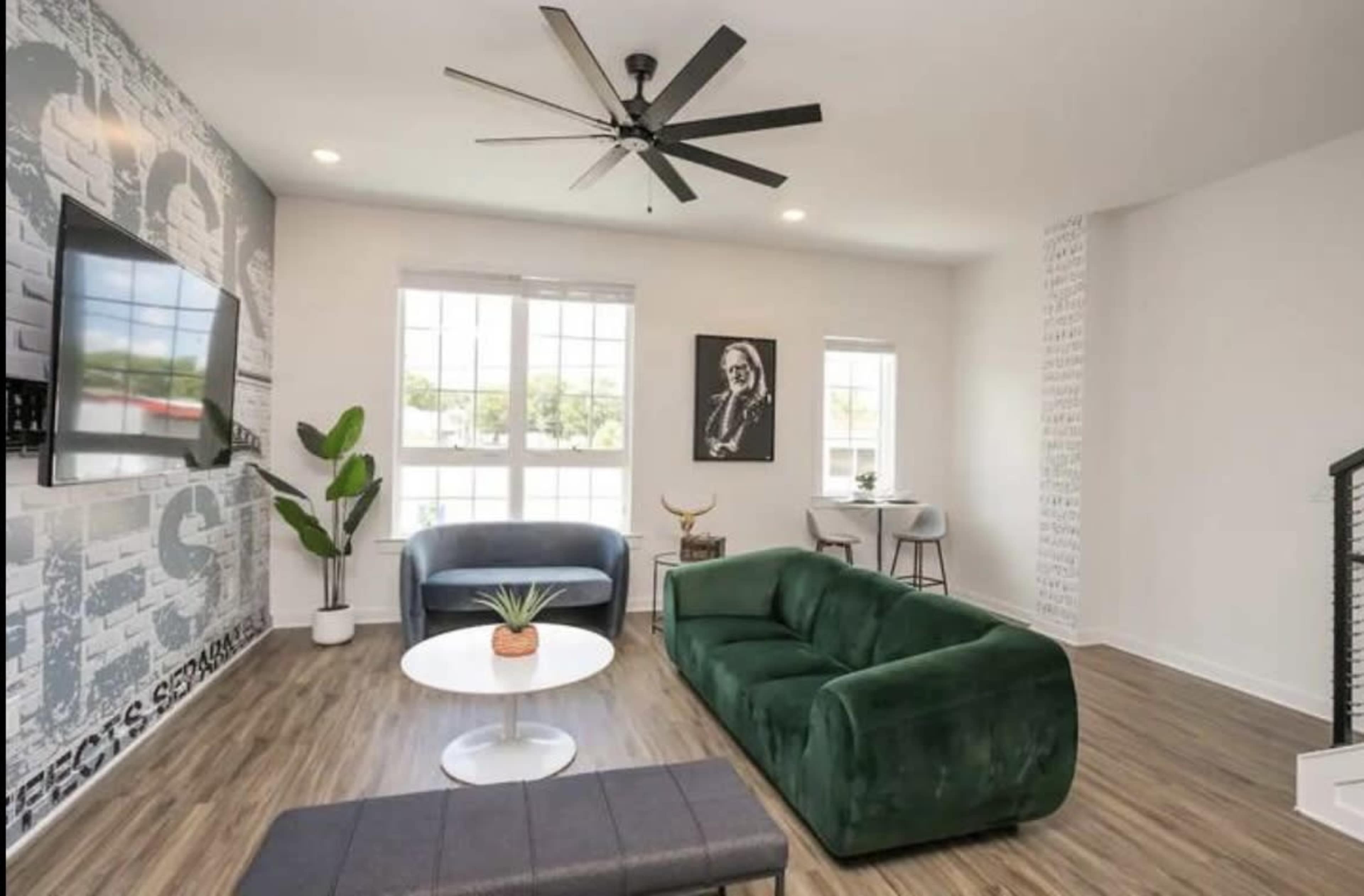 The living room features a green sofa, blue chair, a round coffee table with a plant, a large wall-mounted TV, and large windows allowing natural light.