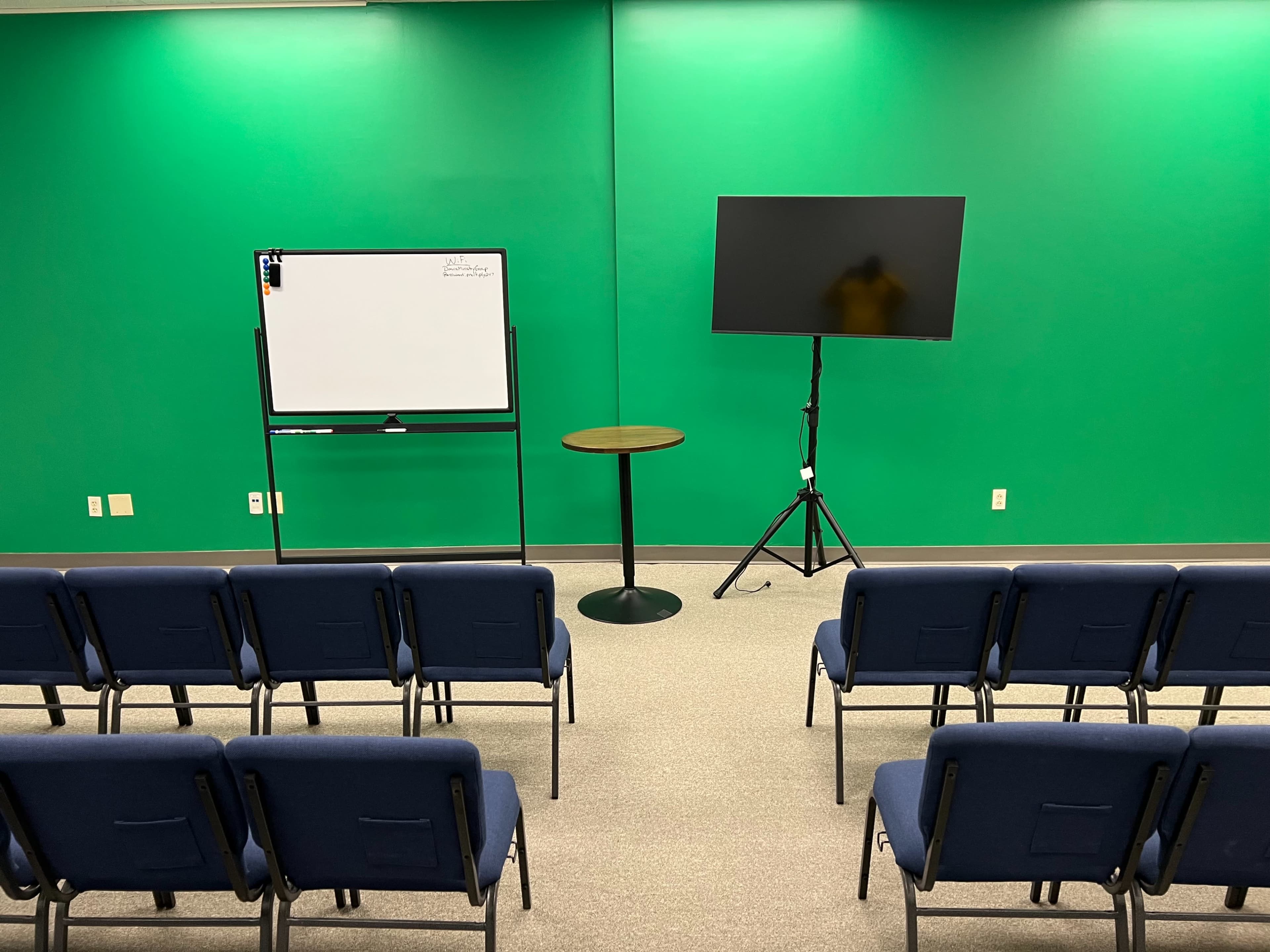 The image shows a room with green walls featuring a whiteboard, a monitor on a stand, and rows of blue chairs arranged facing the presentation area.