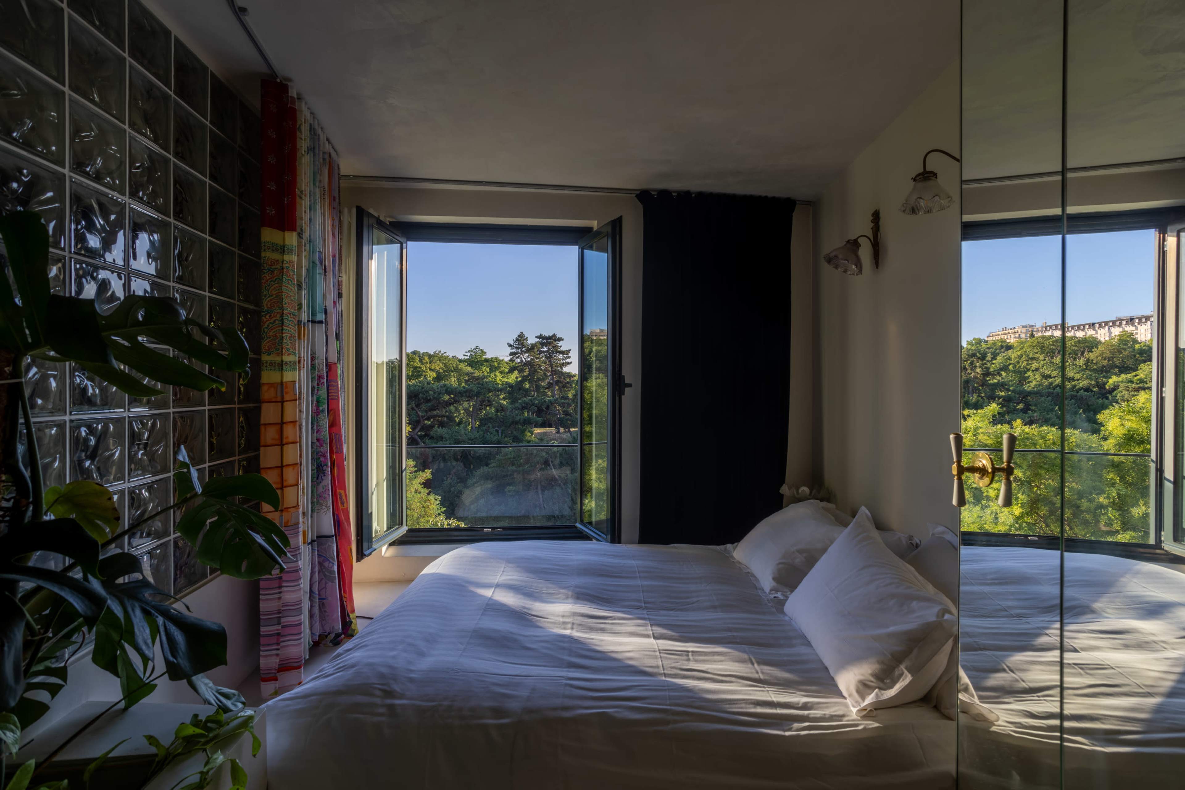 The image shows a bedroom with a large open window overlooking a lush green landscape, featuring a bed with white linens and decorative curtains.