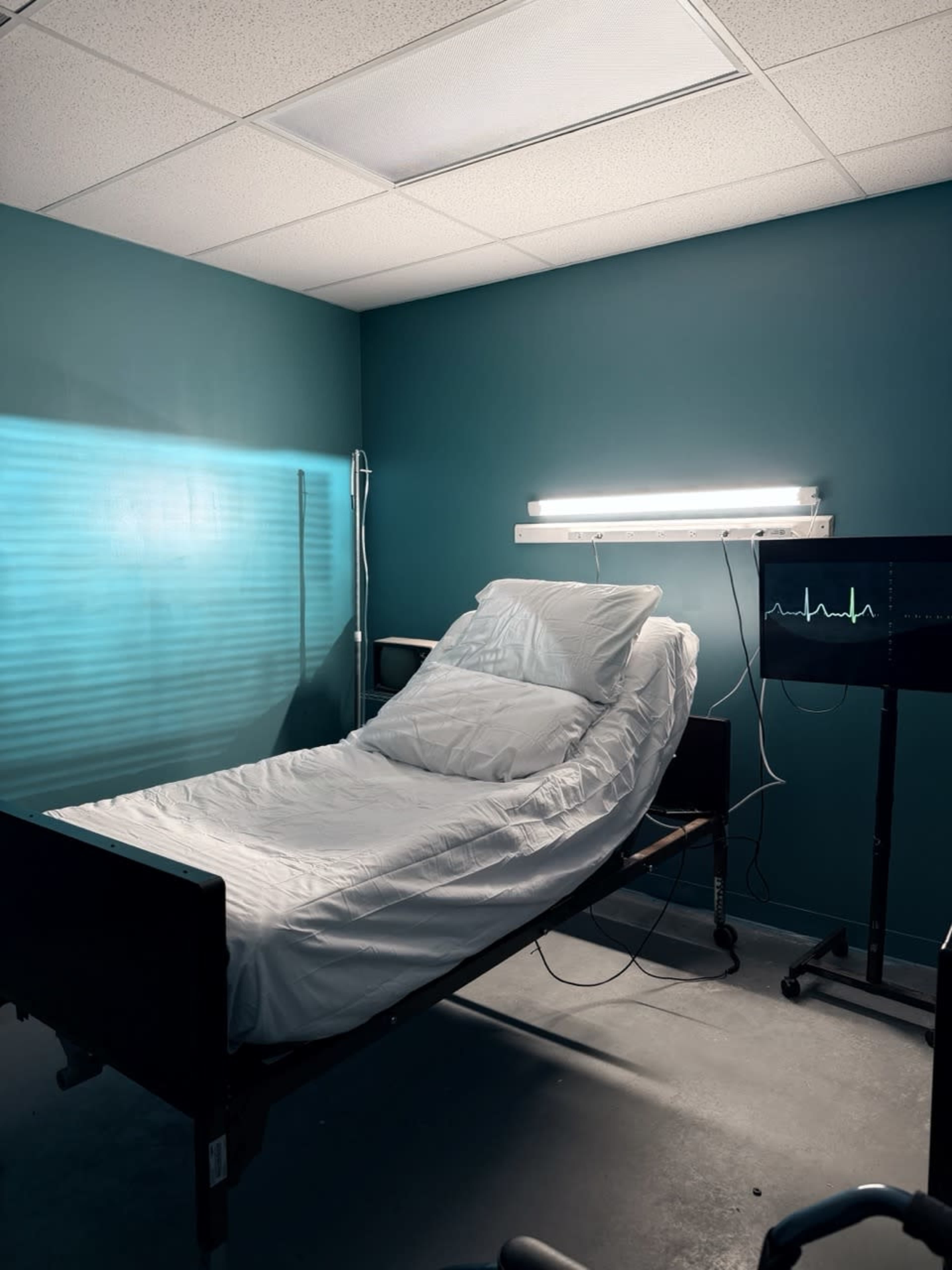 The image shows a hospital room with a single bed made up with white linens and a monitor displaying a health line.
