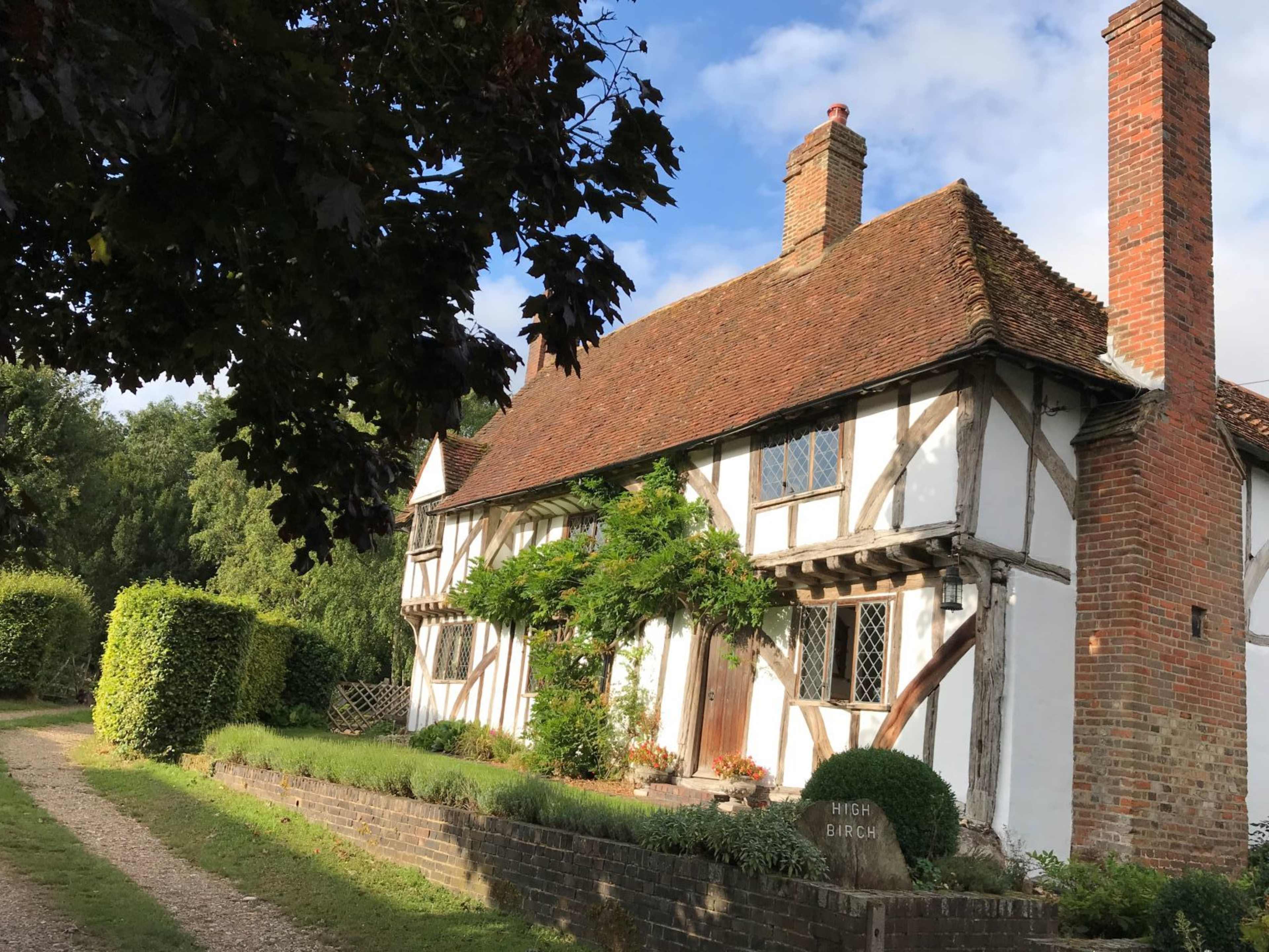 A traditional half-timbered cottage with a thatched roof is situated along a gravel pathway, surrounded by lush greenery.