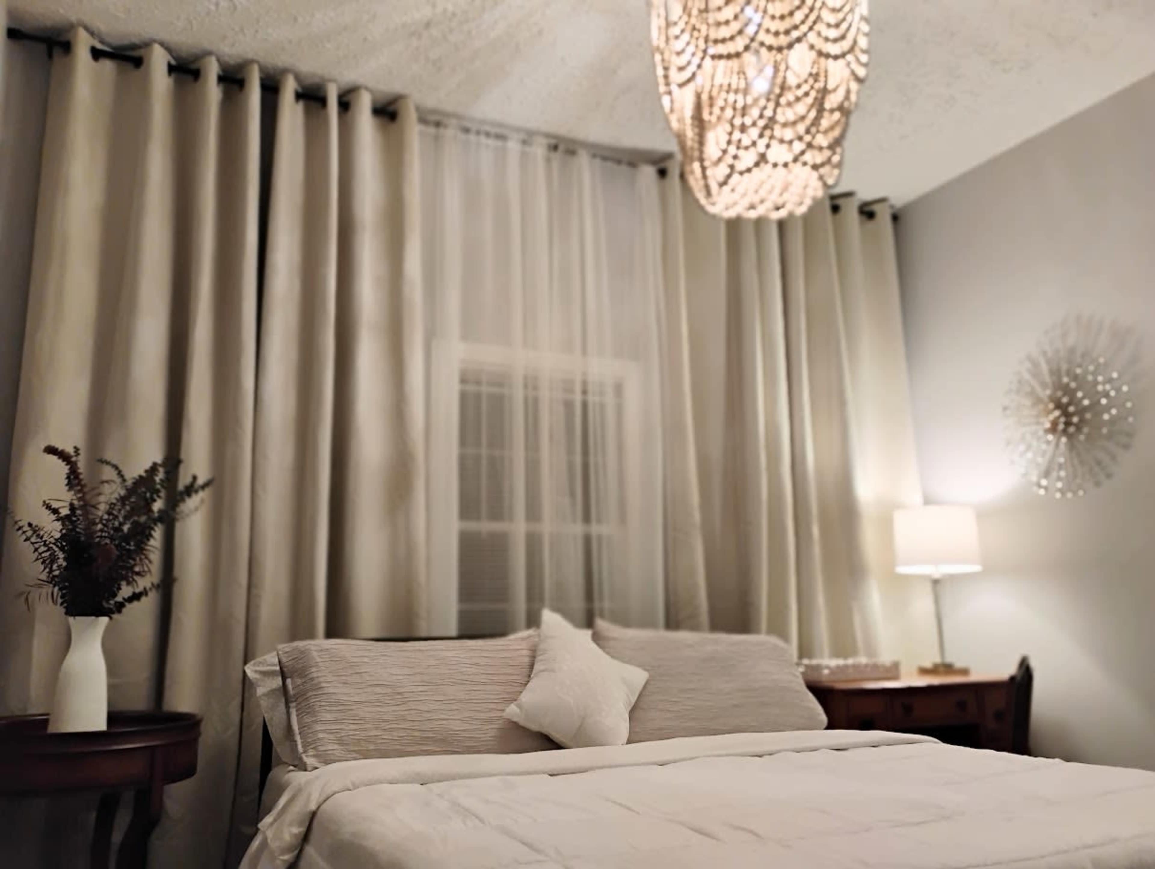A neatly made bed with a decorative pillow is positioned in a softly lit bedroom featuring sheer curtains, a chandelier, and a table lamp.