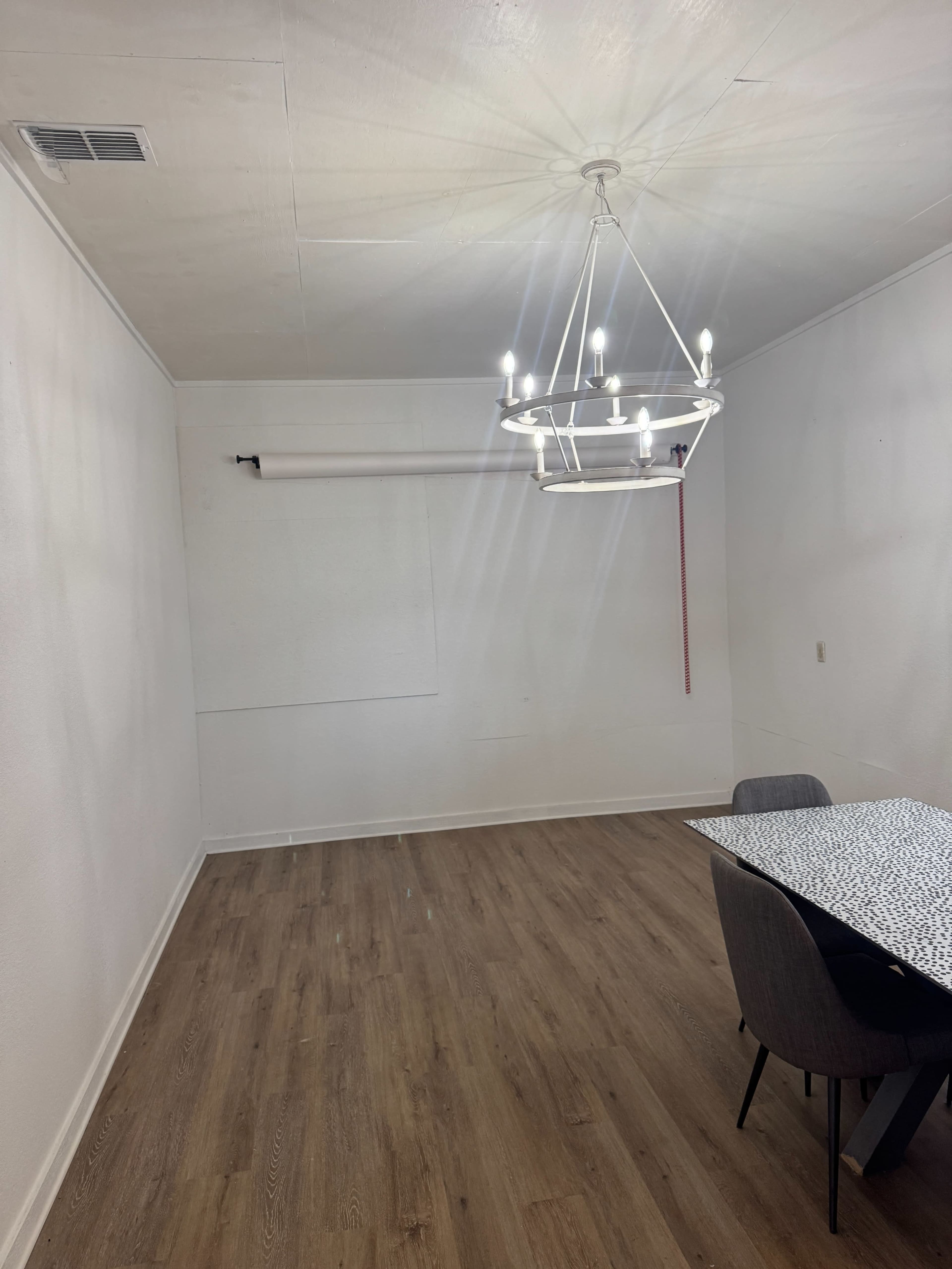 The image shows a sparsely furnished room with light-colored walls, a wooden floor, a modern chandelier, and a table with gray chairs.
