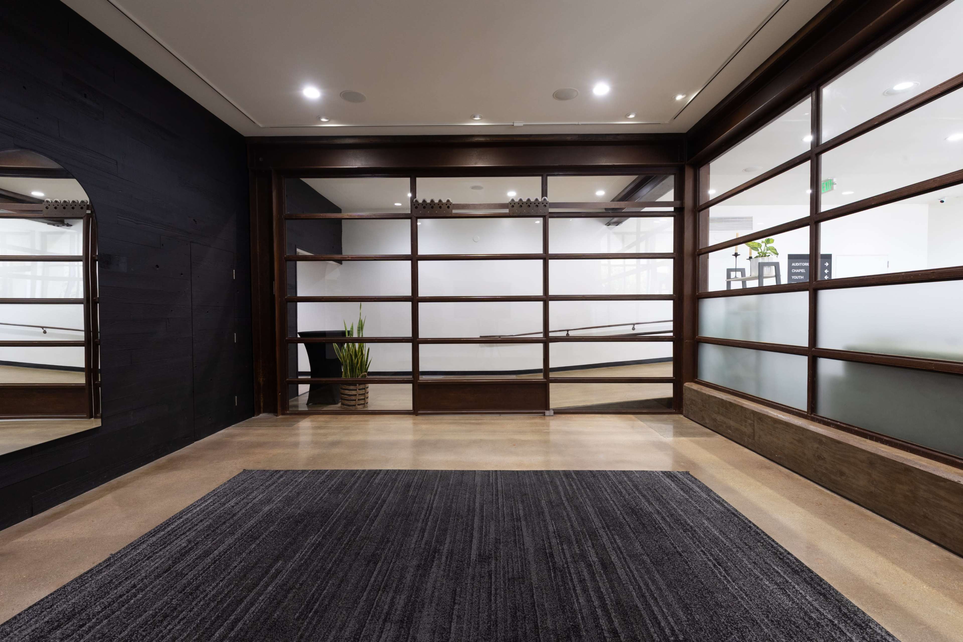The image shows a modern interior space featuring large glass panels, wooden framing, and a dark carpet on a polished concrete floor.