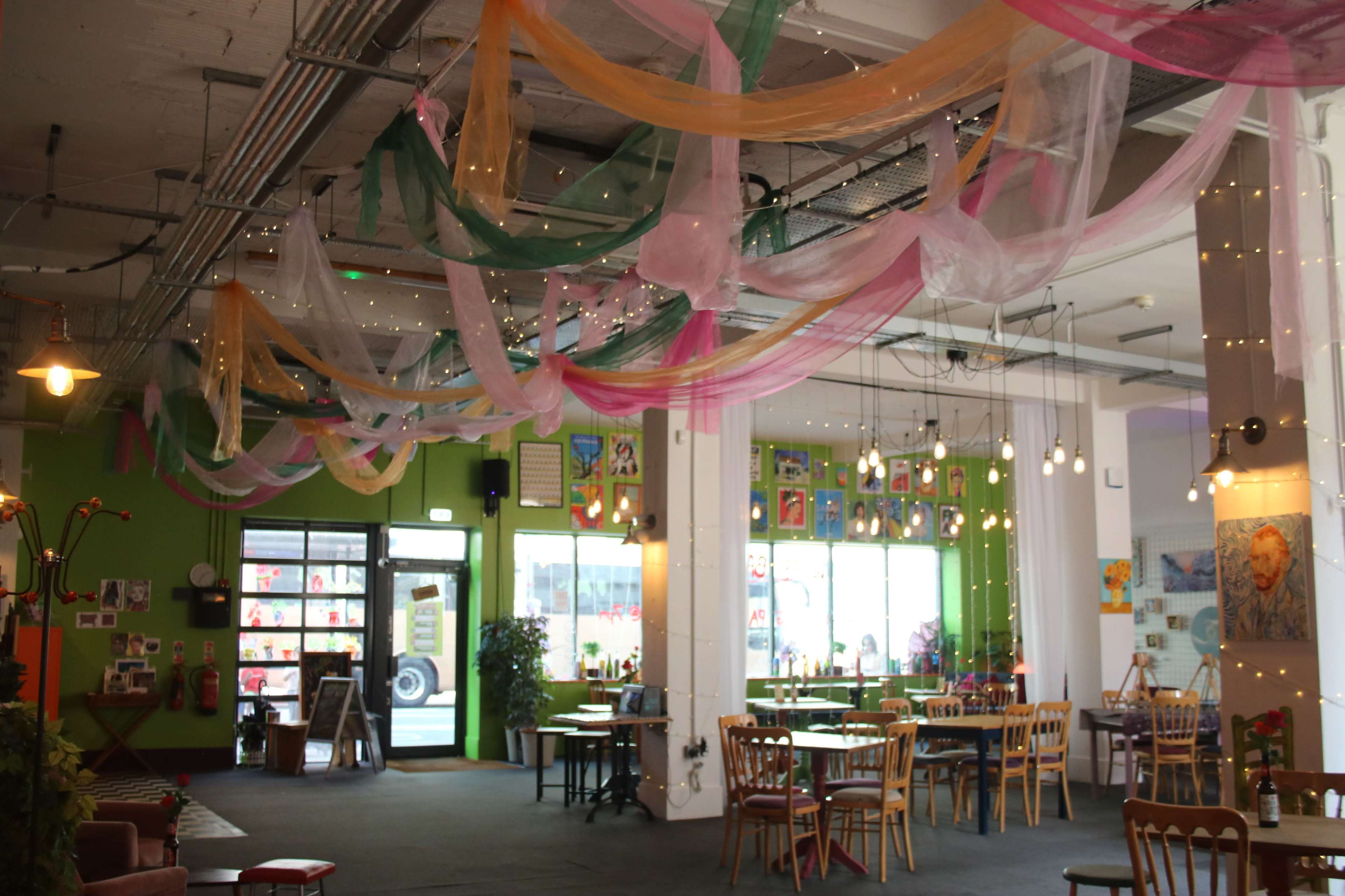 The image shows a spacious indoor café with colorful fabric draping overhead, a mix of tables and chairs, and various artworks displayed on the walls.