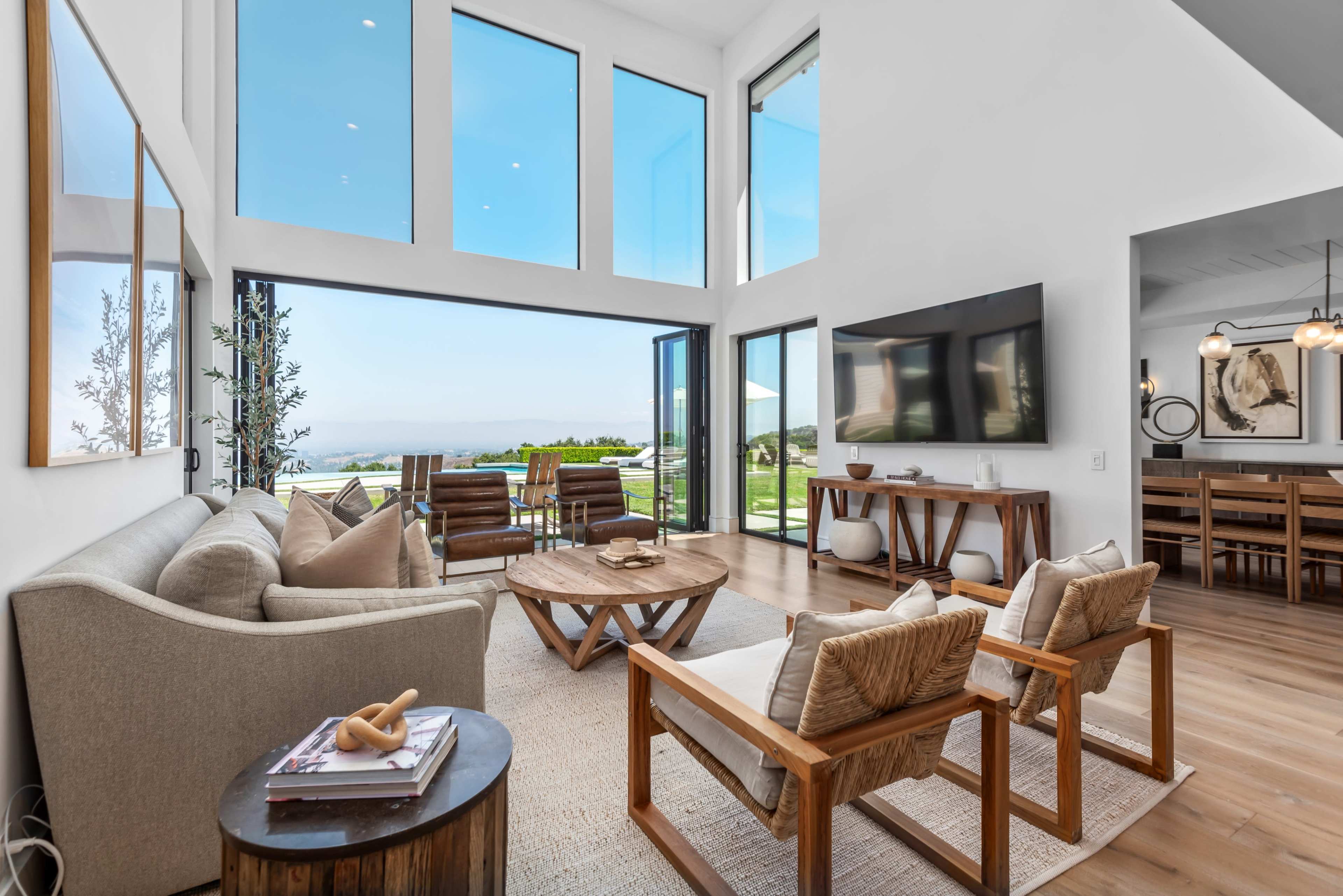 A bright living room features large windows overlooking a scenic landscape, with a cozy seating area centered around a circular wooden coffee table.