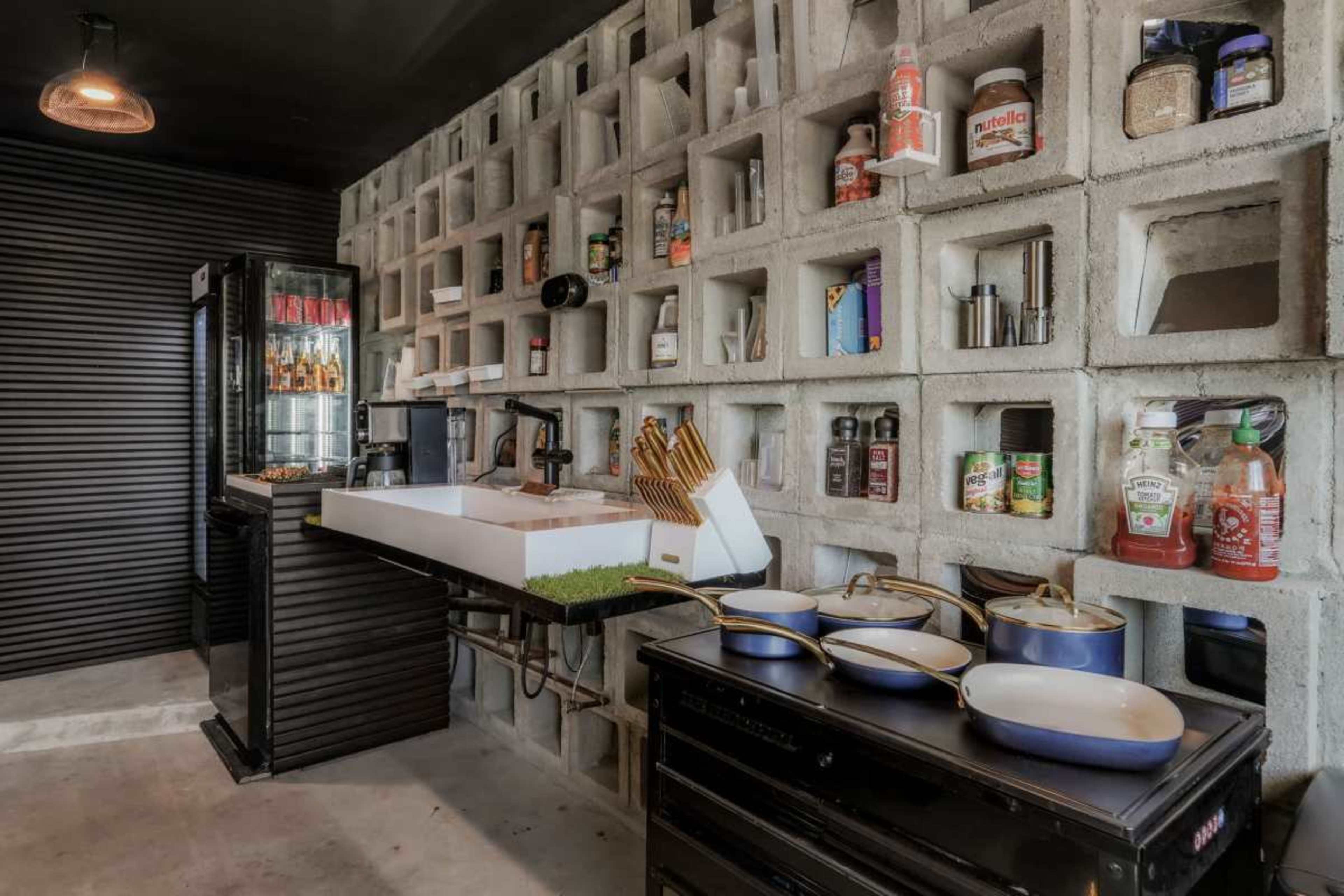 The image shows a modern kitchen with a concrete block wall, an island with a sink and cutting board, and various kitchen items displayed on shelves above and below.