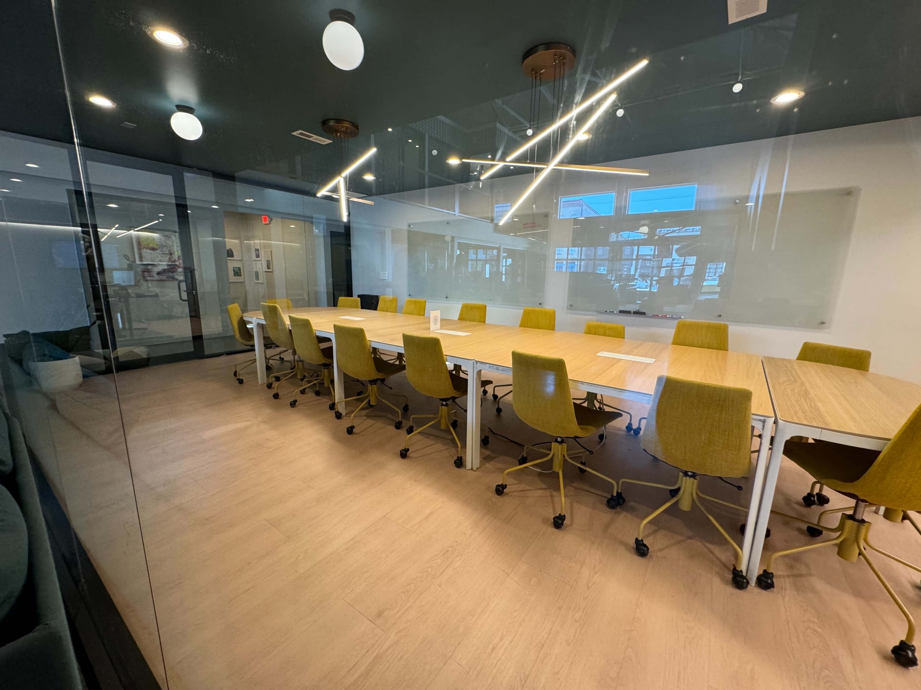 A large, modern conference room features a long wooden table surrounded by yellow office chairs, with large windows allowing natural light to fill the space.