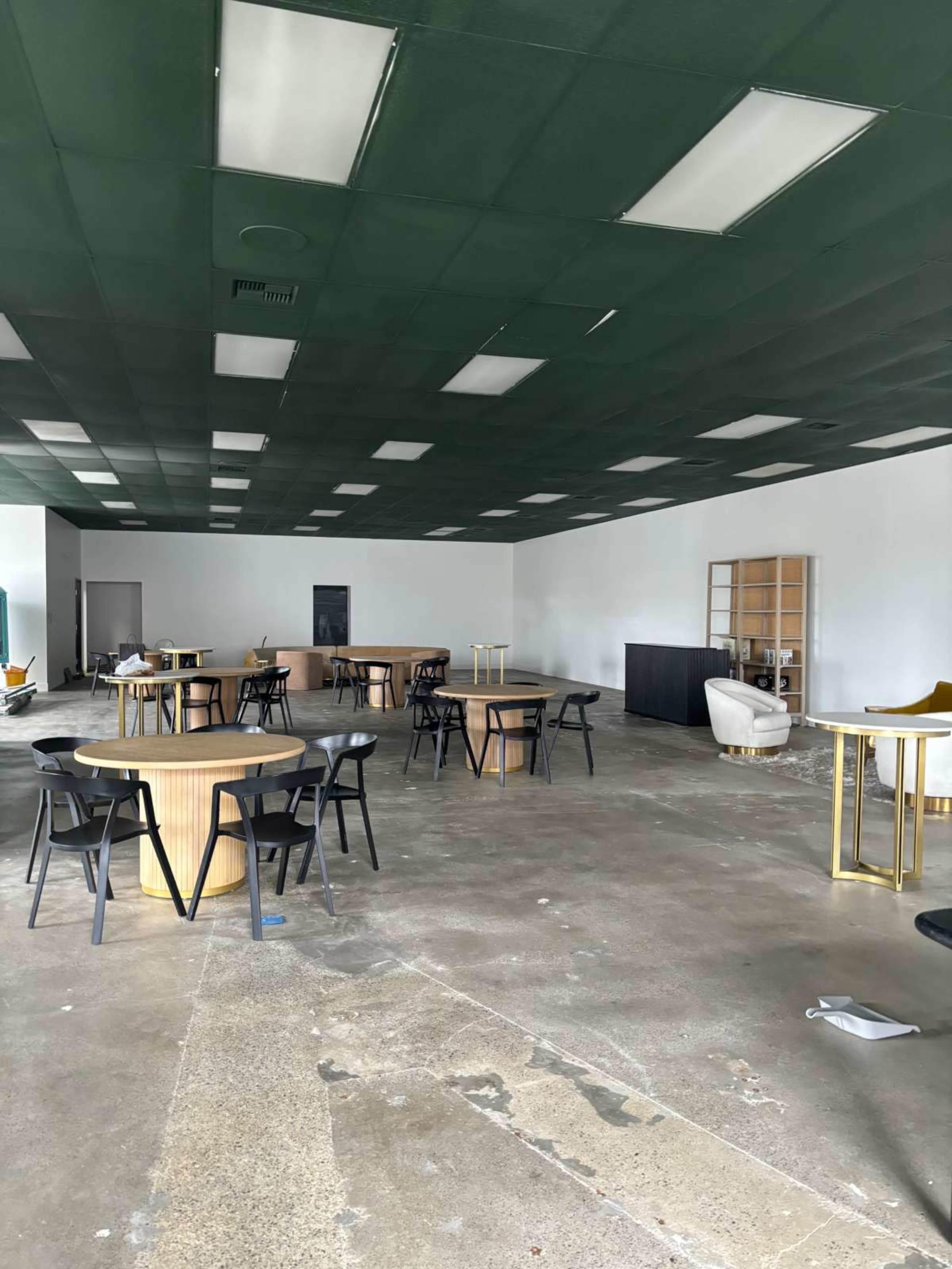 The image shows a spacious, empty room with several round tables and chairs scattered throughout, featuring a green ceiling and unfinished concrete flooring.