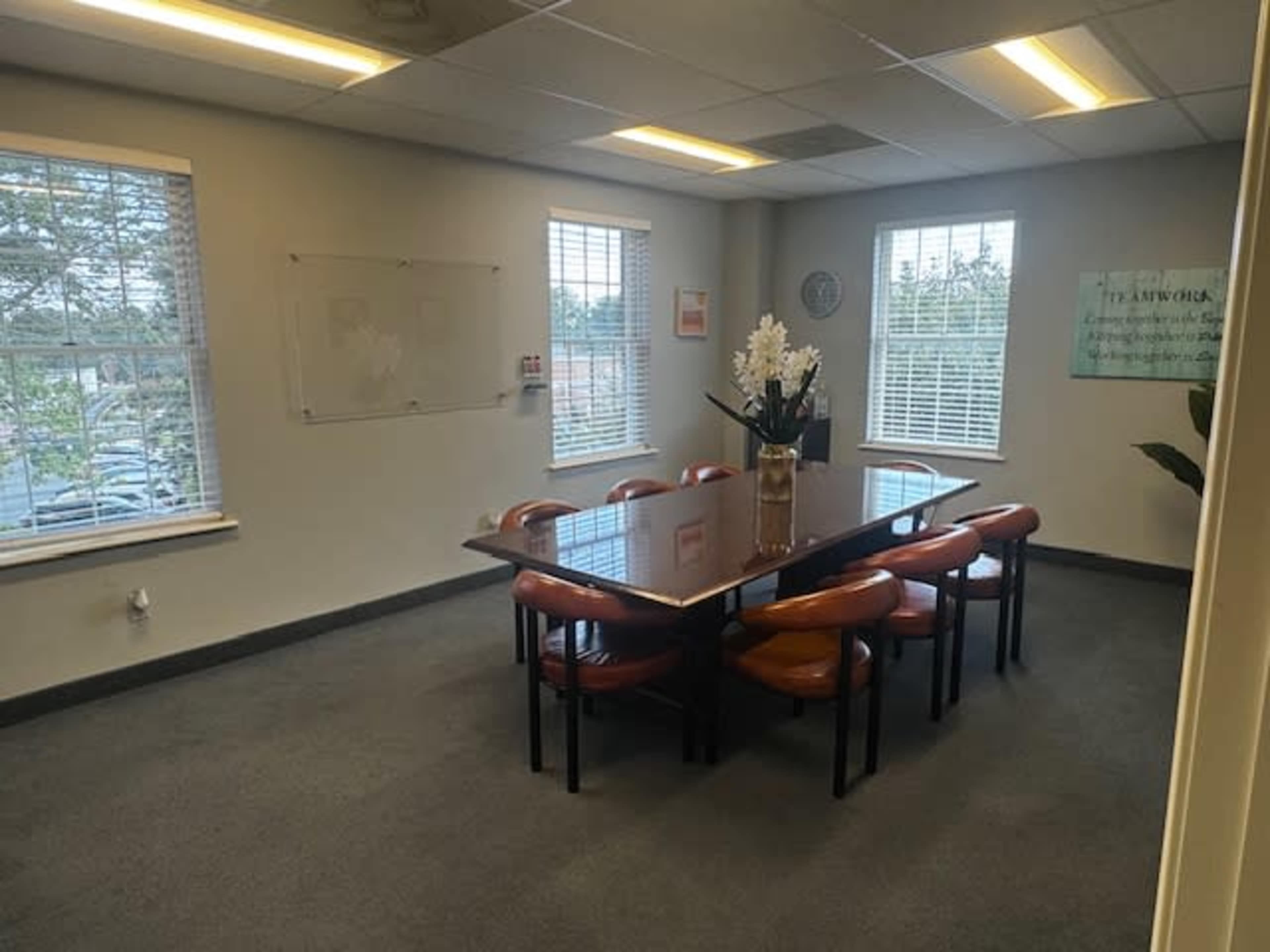 A meeting room features a large table surrounded by several chairs, with windows allowing natural light and a whiteboard on the wall.