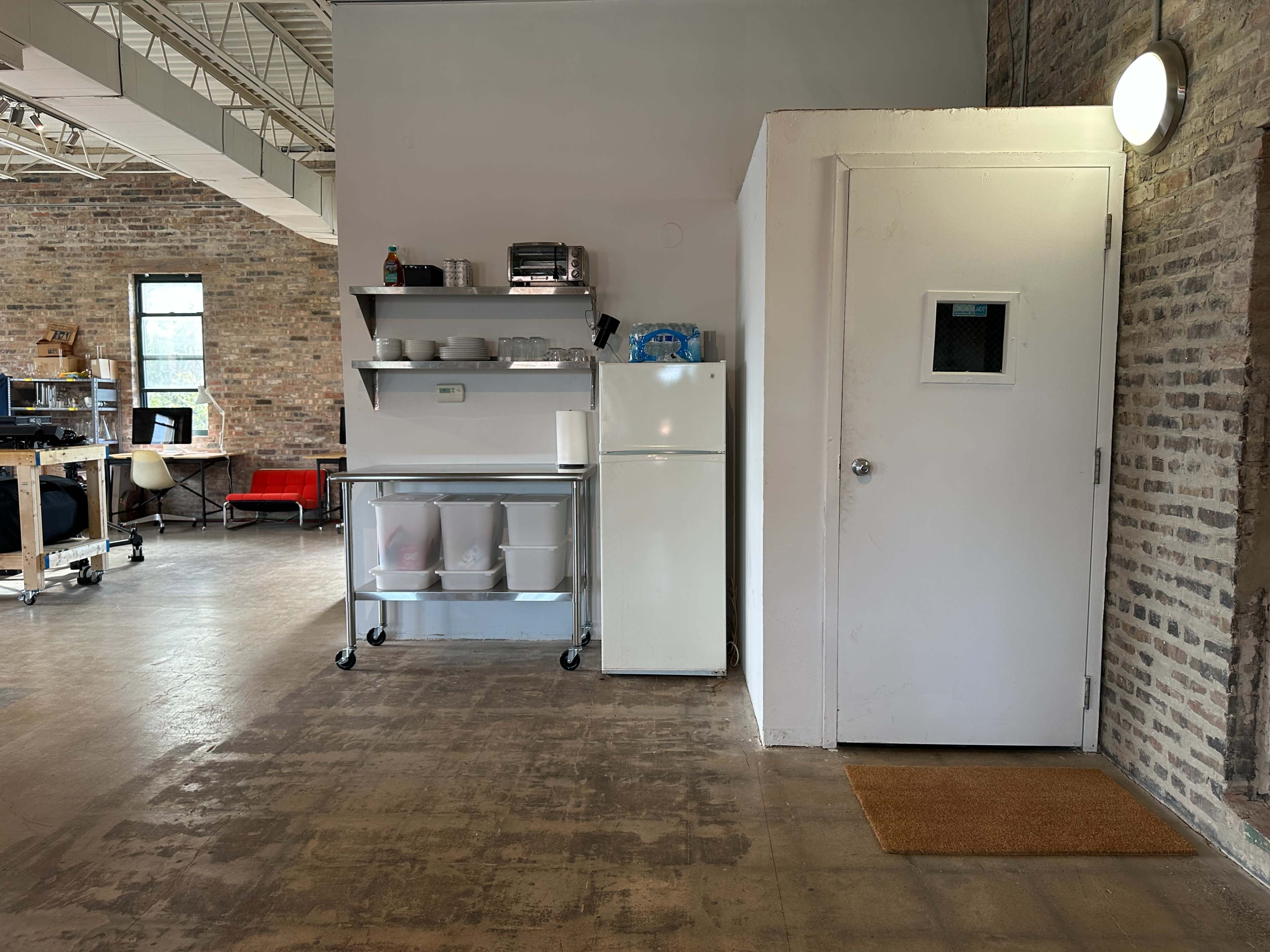 The image shows a room with exposed brick walls featuring a kitchen area with a refrigerator, shelves, and a closed door with a small window.