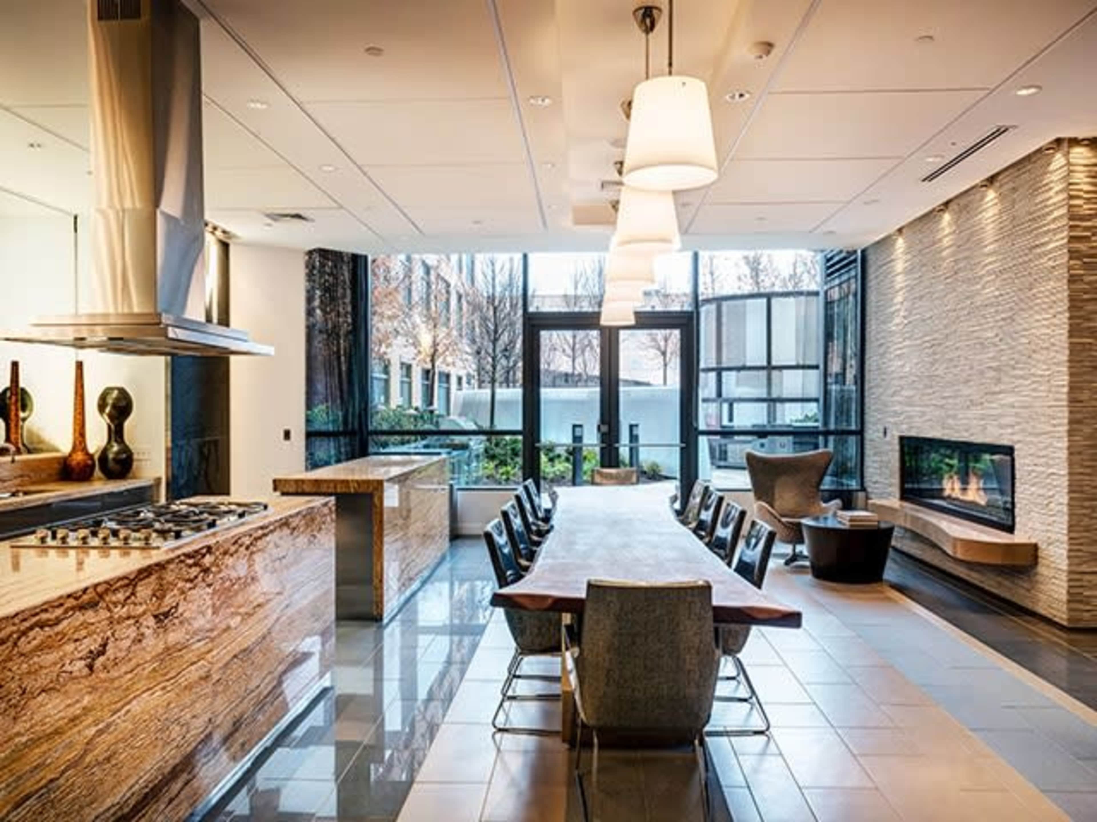 A modern kitchen and dining area features a large wooden table surrounded by sleek chairs, with a window offering a view of an outdoor space, and a fireplace integrated into the wall.