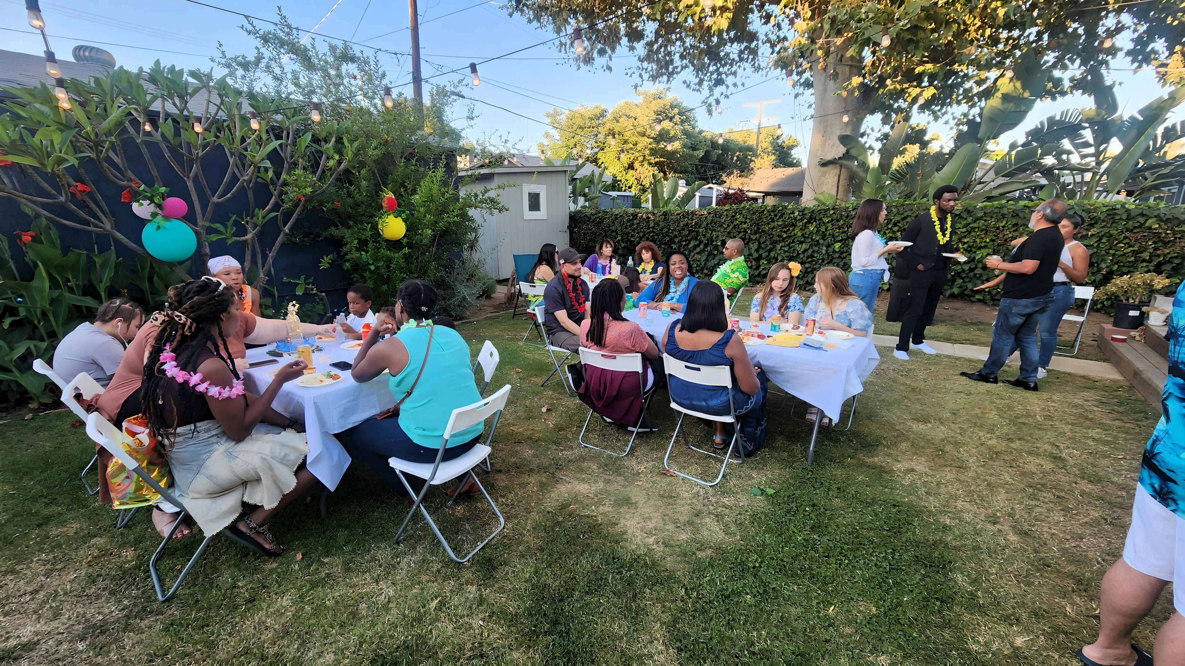 A group of people is seated at tables outside in a backyard, enjoying a meal and celebrating together.