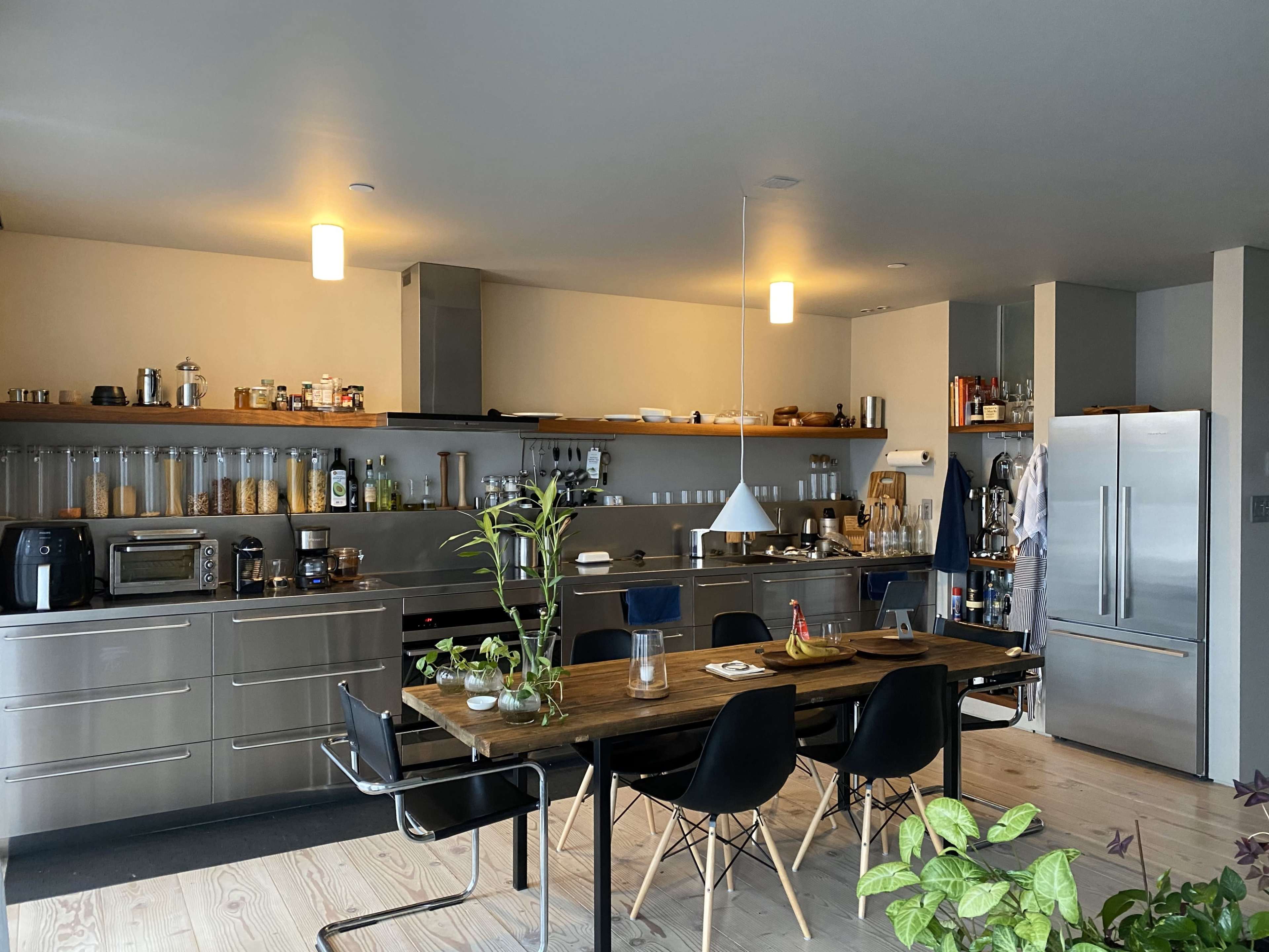A modern kitchen featuring stainless steel appliances, a central dining table, and shelves stocked with various containers and kitchen items.