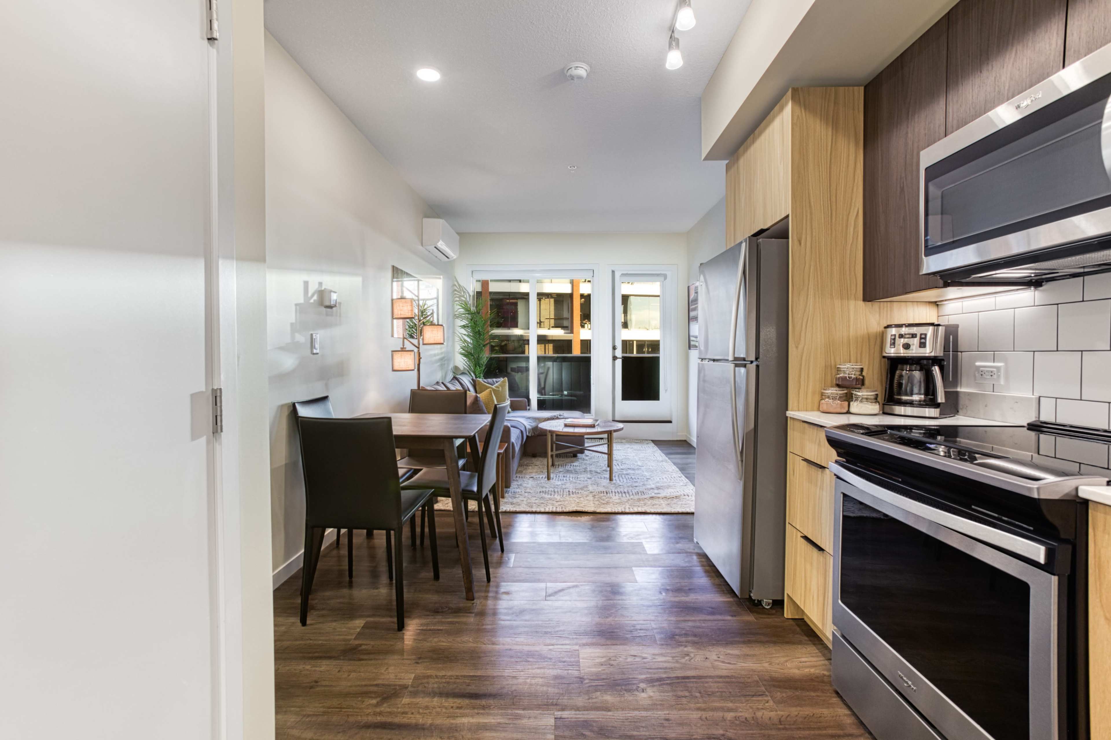 The image shows a modern kitchen and dining area with a sleek design, featuring stainless steel appliances, wooden cabinetry, a dining table with chairs, and a living space with a sofa and large windows.