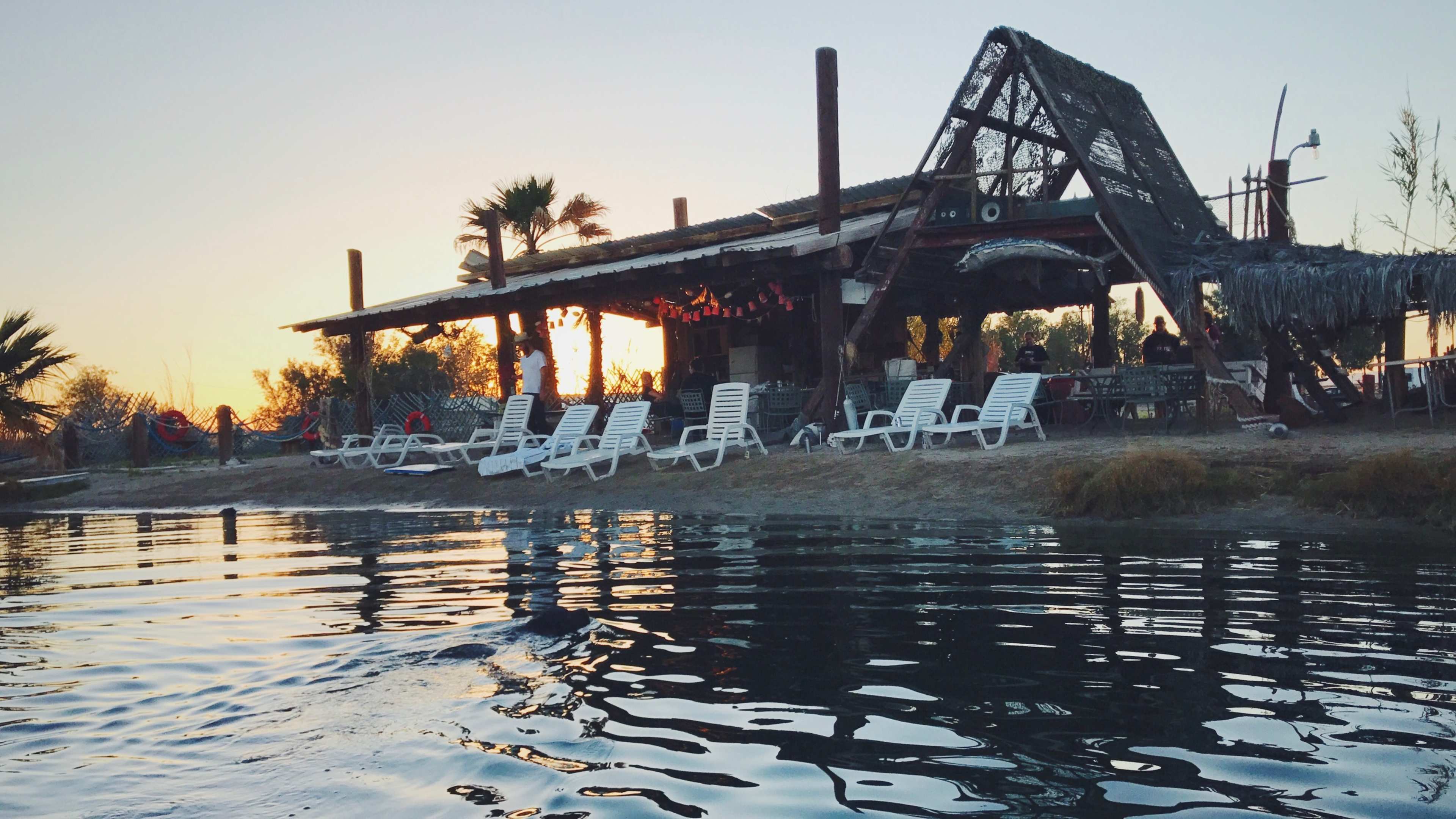 A thatched-roof structure stands by the water, with several white lounge chairs lined up on the sandy shore during sunset.