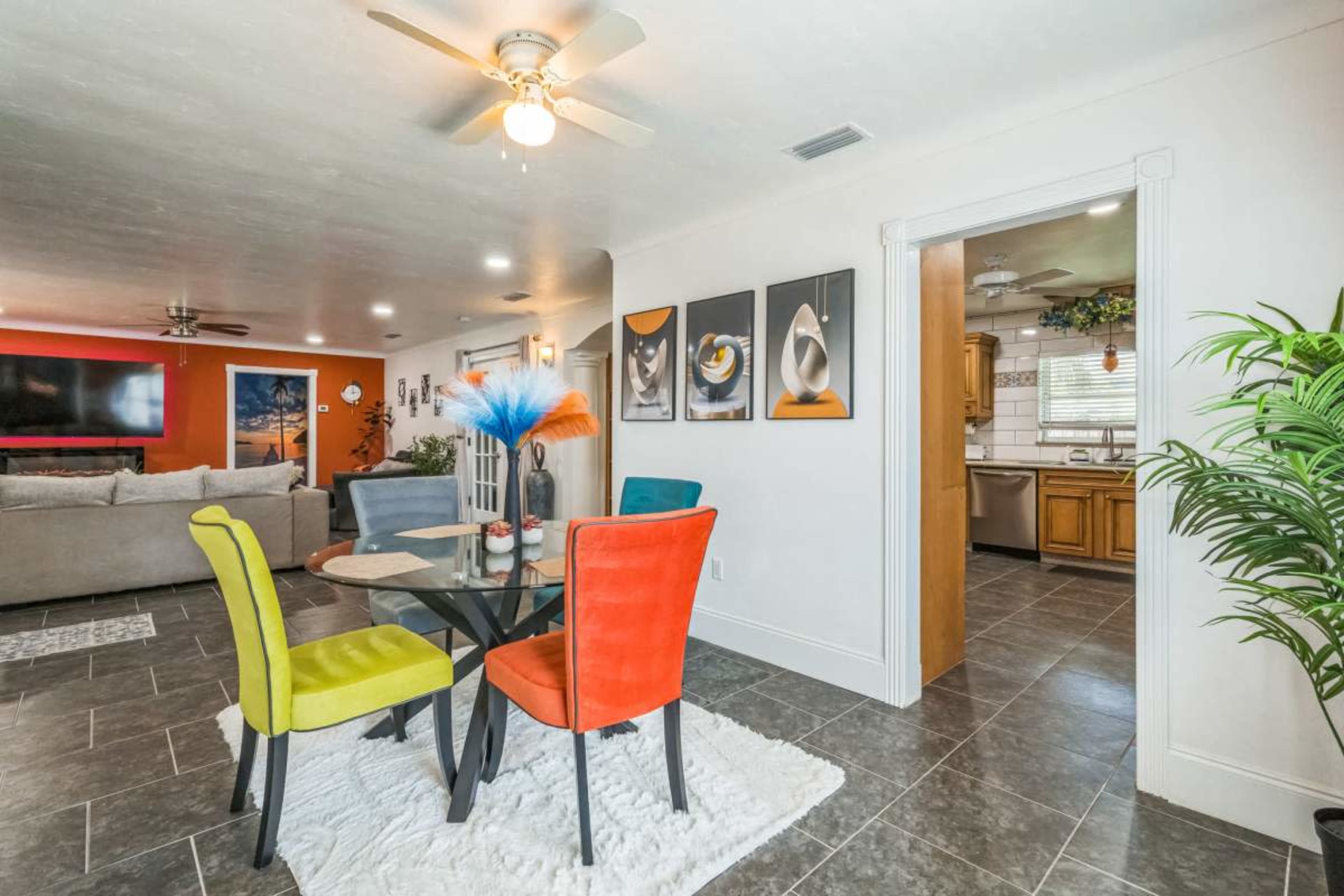 The image features a modern dining area with a round glass table surrounded by colorful chairs, set against a backdrop of a living room with a gray sofa and vibrant wall accents.