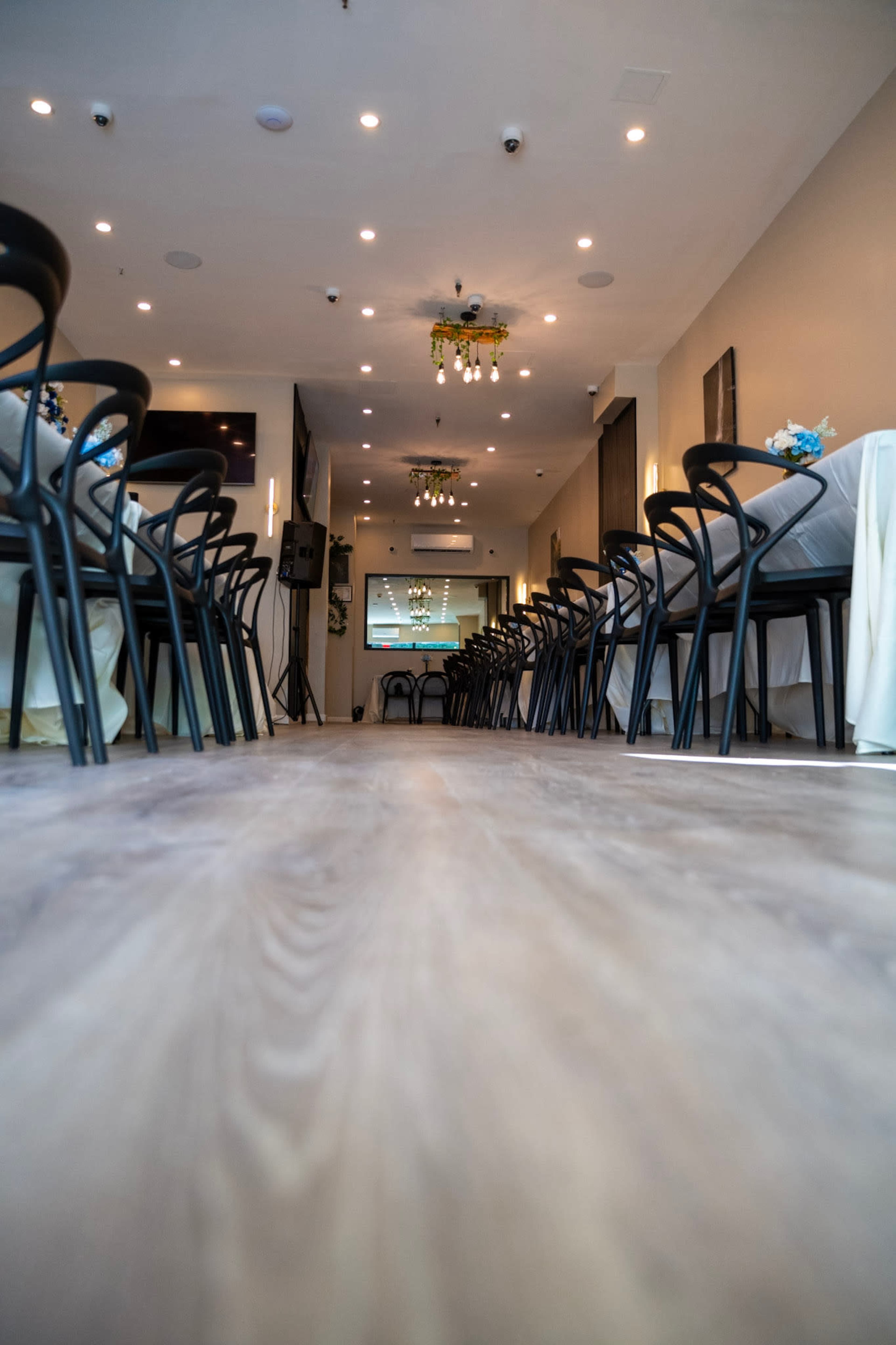 A narrow interior view of a restaurant featuring rows of tables with white tablecloths and dark chairs, illuminated by overhead lights.
