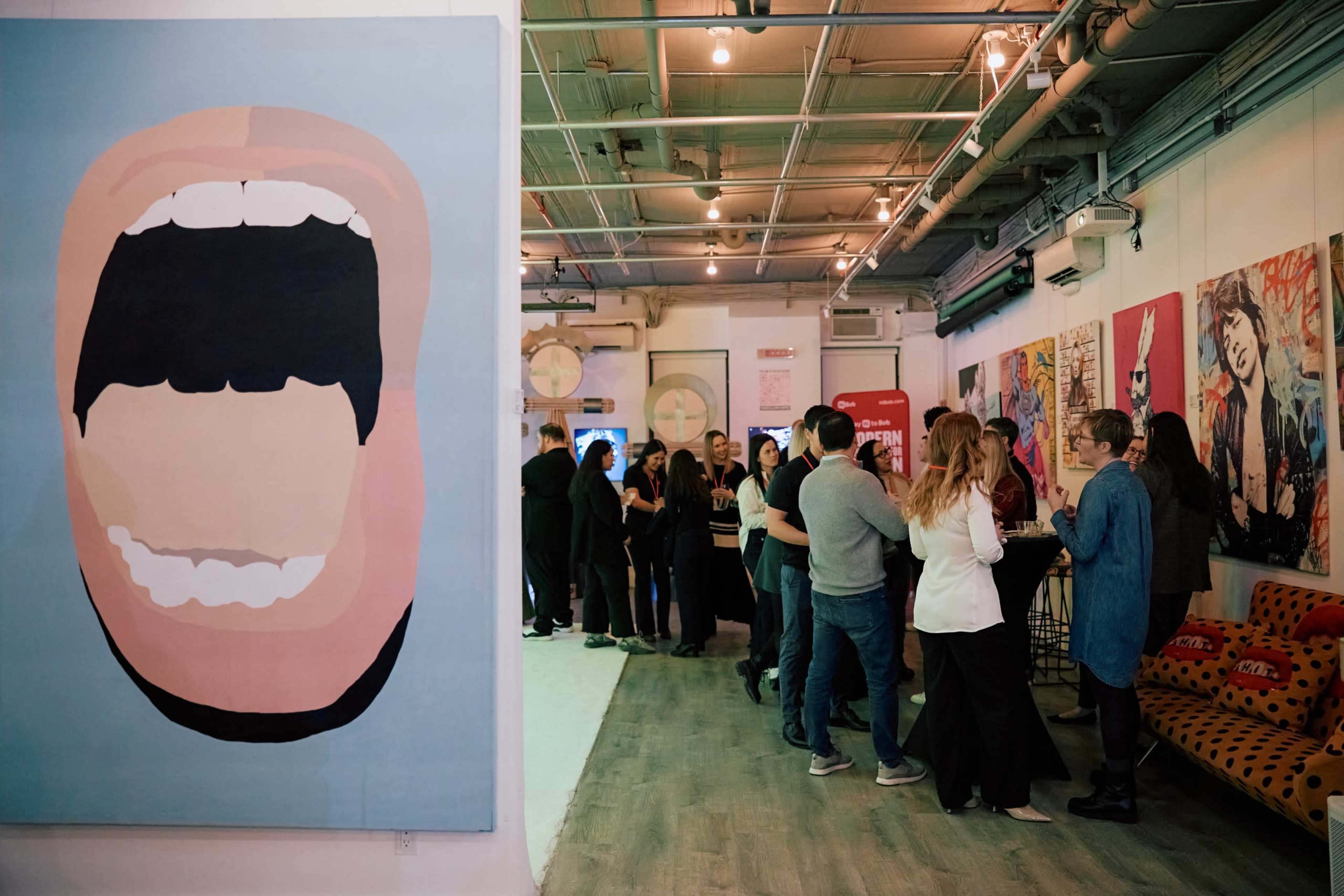 A large abstract painting of an open mouth is positioned on the left side of a gallery where people are mingling and socializing, surrounded by colorful artwork on the walls.