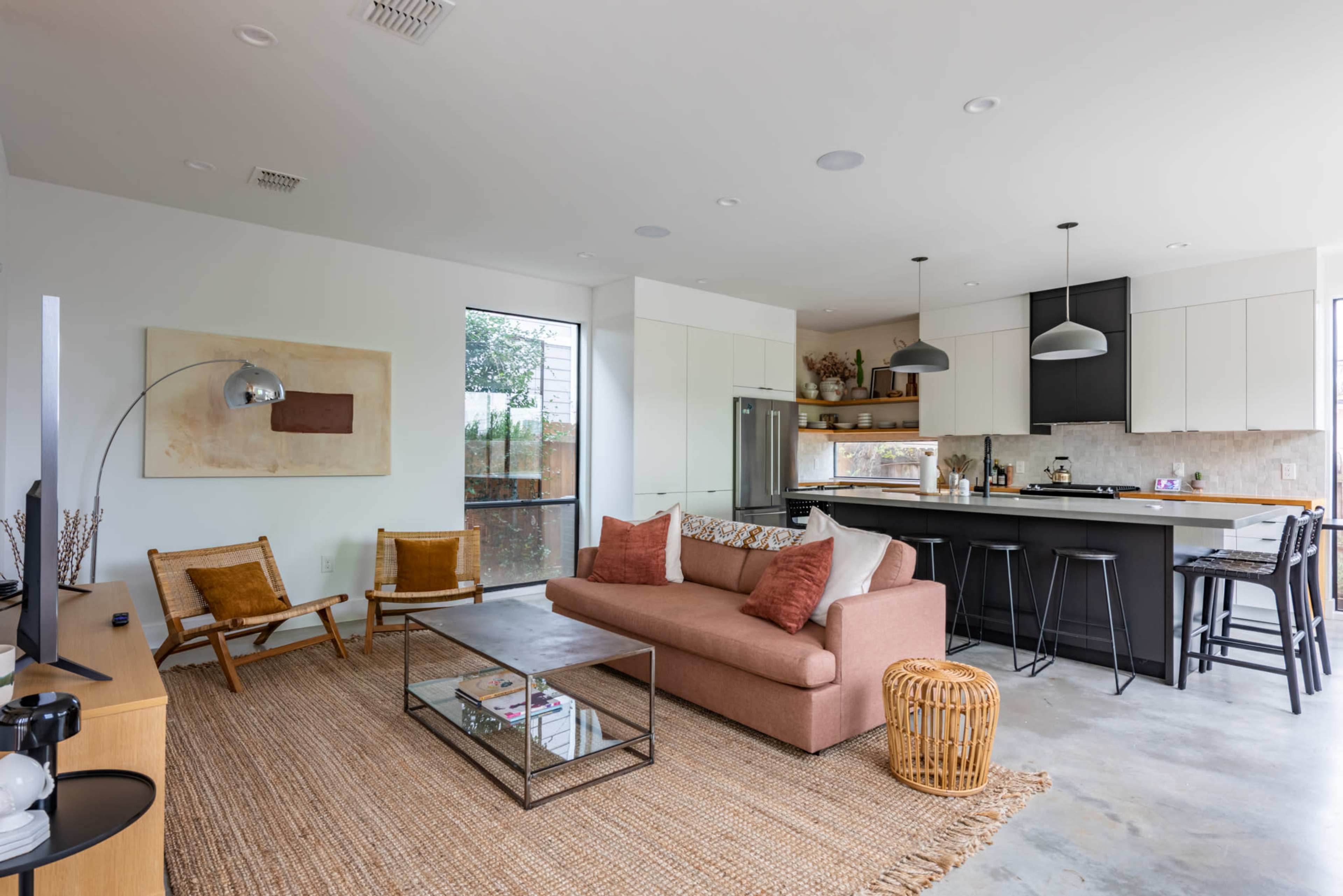 A modern living room features a pink sofa, a glass coffee table, and wooden furniture, with an open kitchen visible in the background.