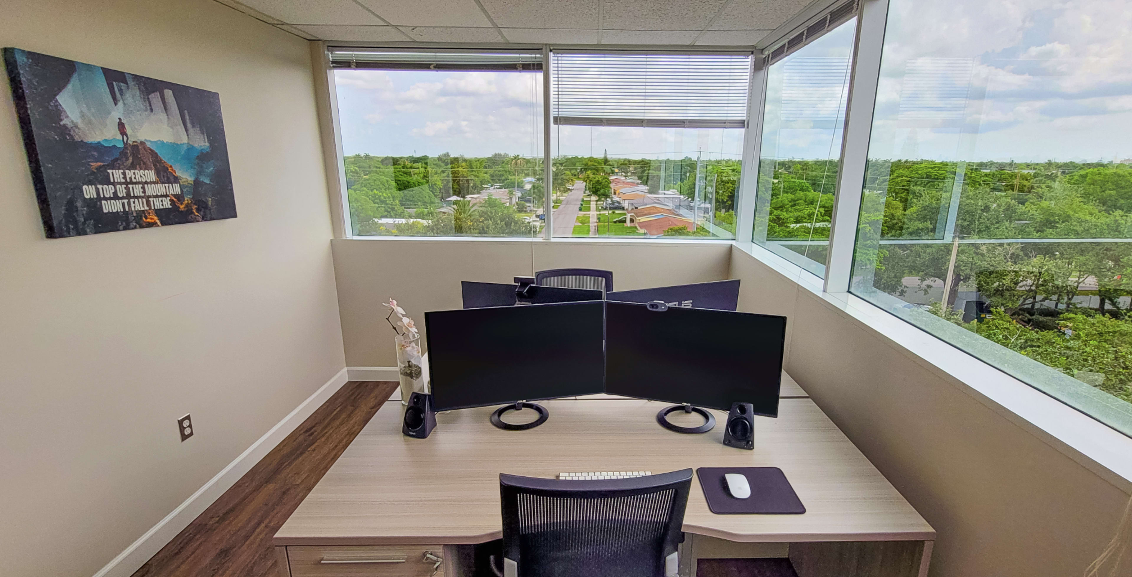 A modern office workspace featuring dual monitors on a desk, with a view of greenery and streets visible through large windows.
