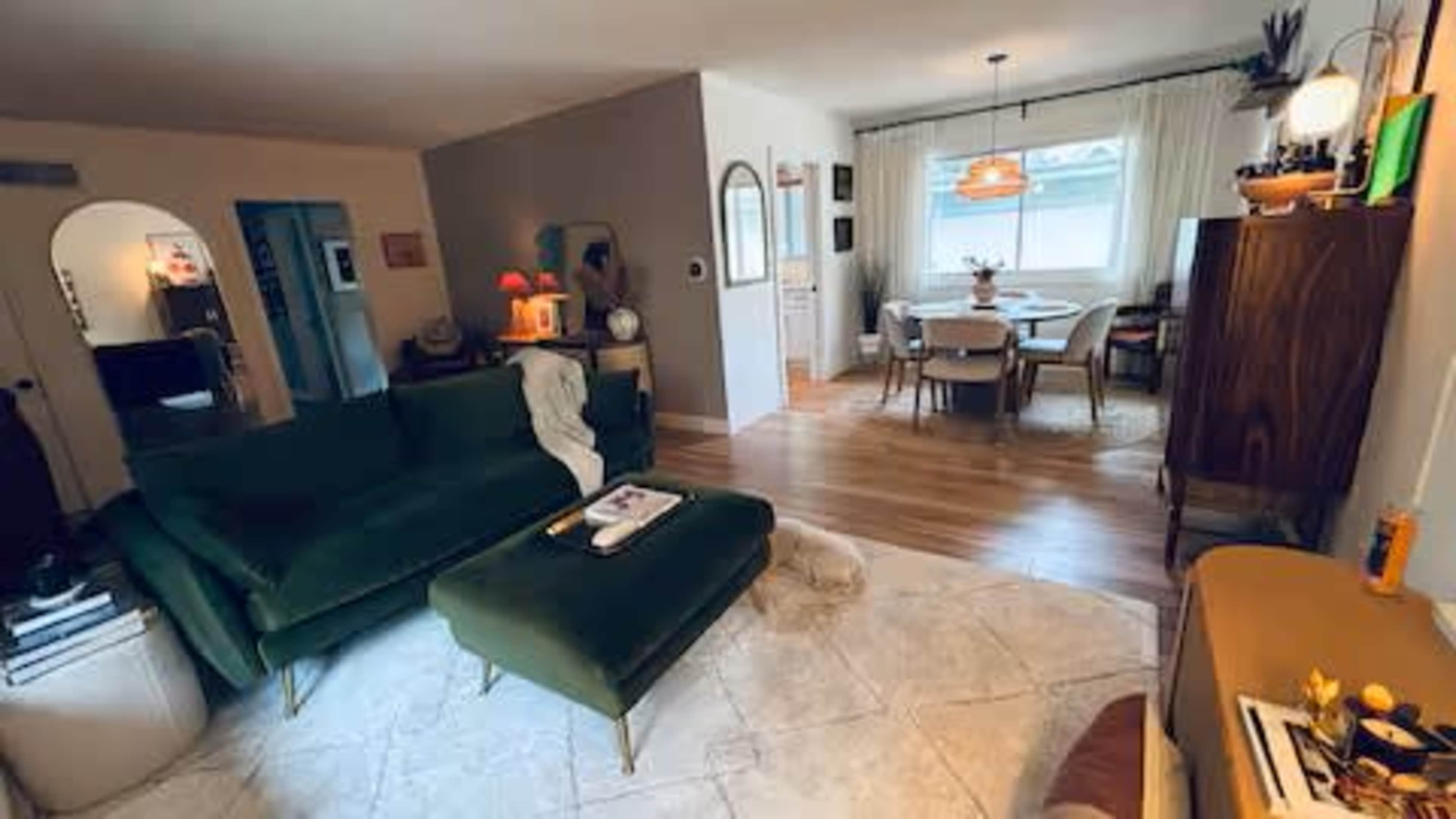 The image shows a cozy living room with a green sofa and ottoman, leading into a dining area with a round table and several chairs.