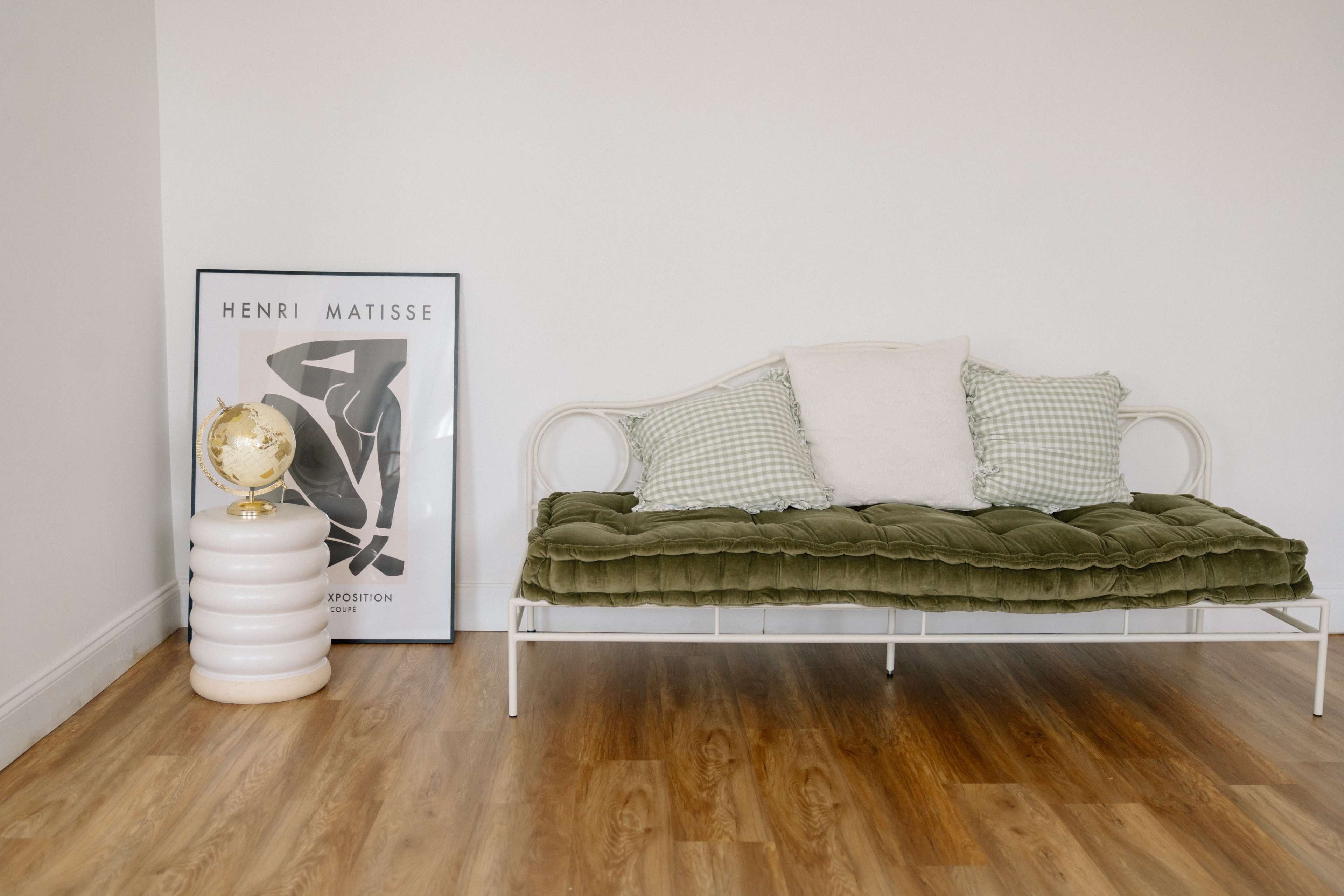 The image shows a minimalist seating area with a green velvet sofa, a framed artwork by Henri Matisse, and a white side table with a decorative globe in the corner.