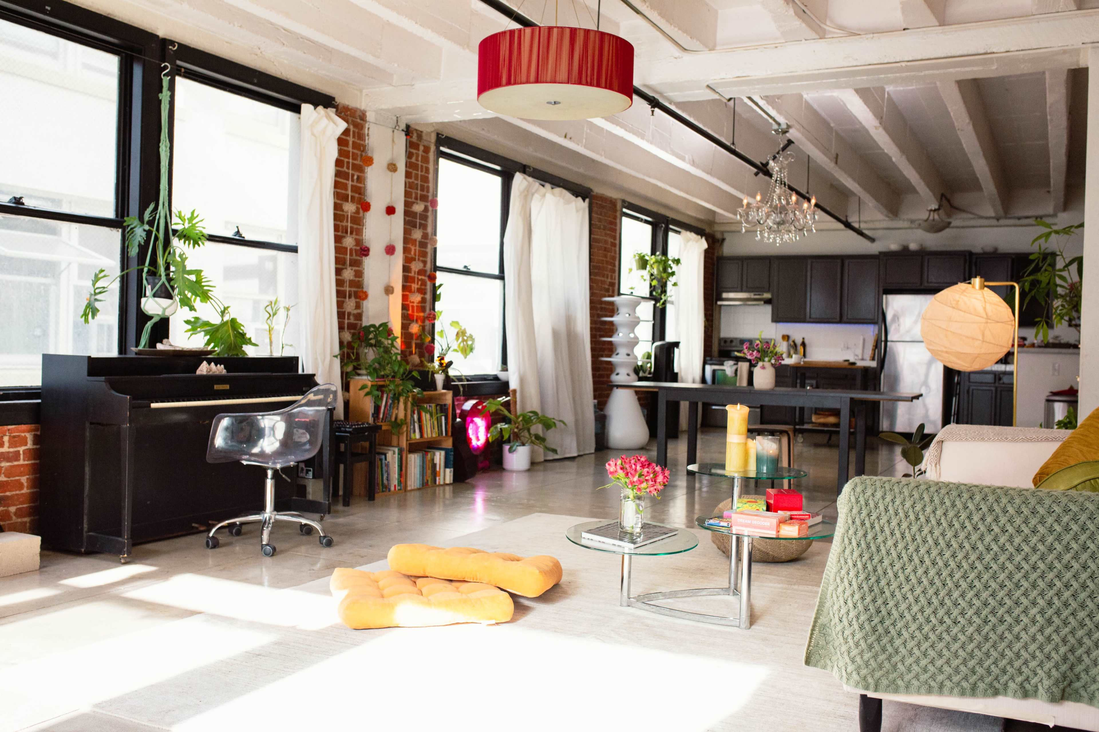 A bright, spacious loft features large windows, a piano, plants, and a modern seating area with a glass coffee table.