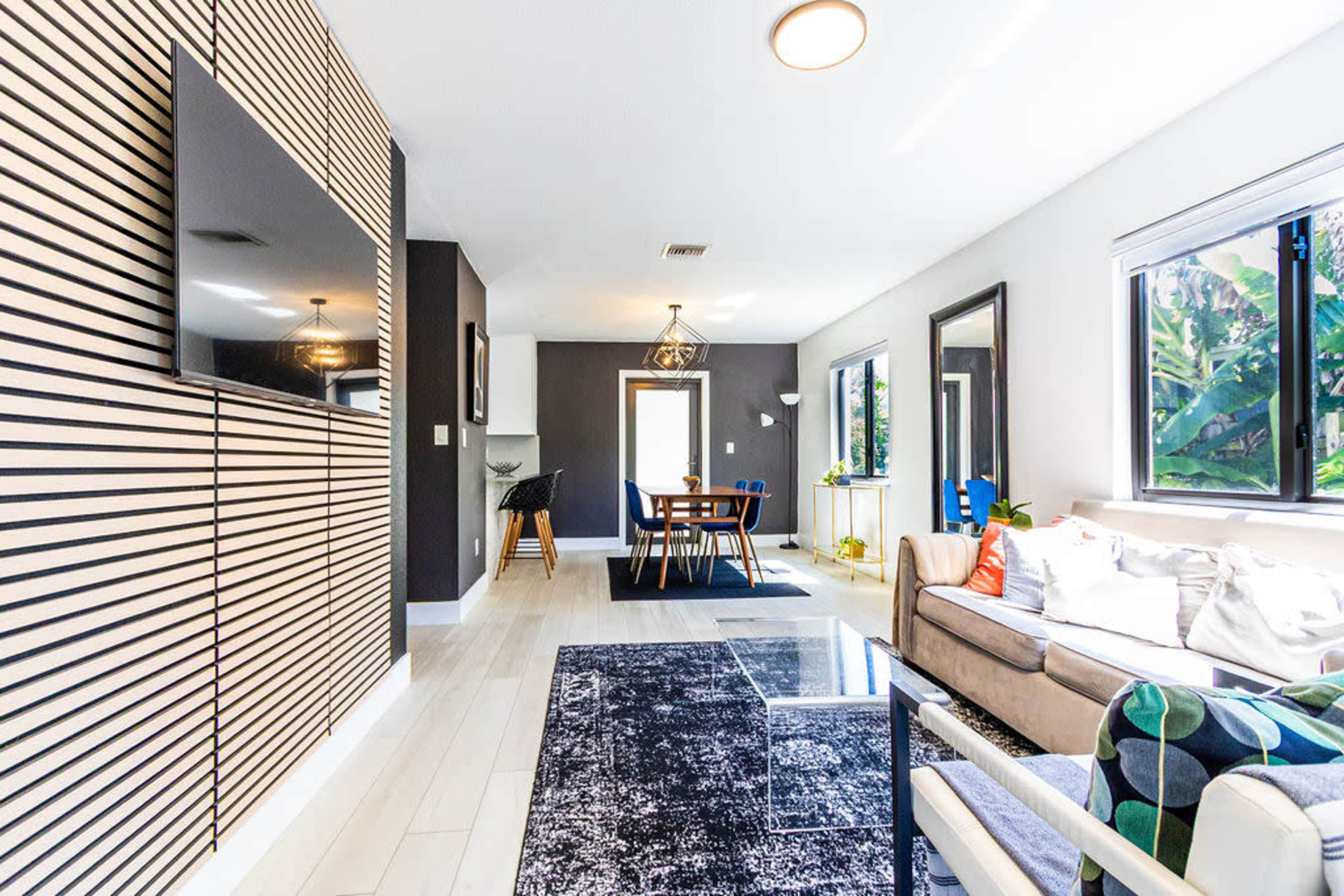 The image shows a modern living room with a television mounted on a striped accent wall, a dining area in the background, and ample natural light coming through the windows.