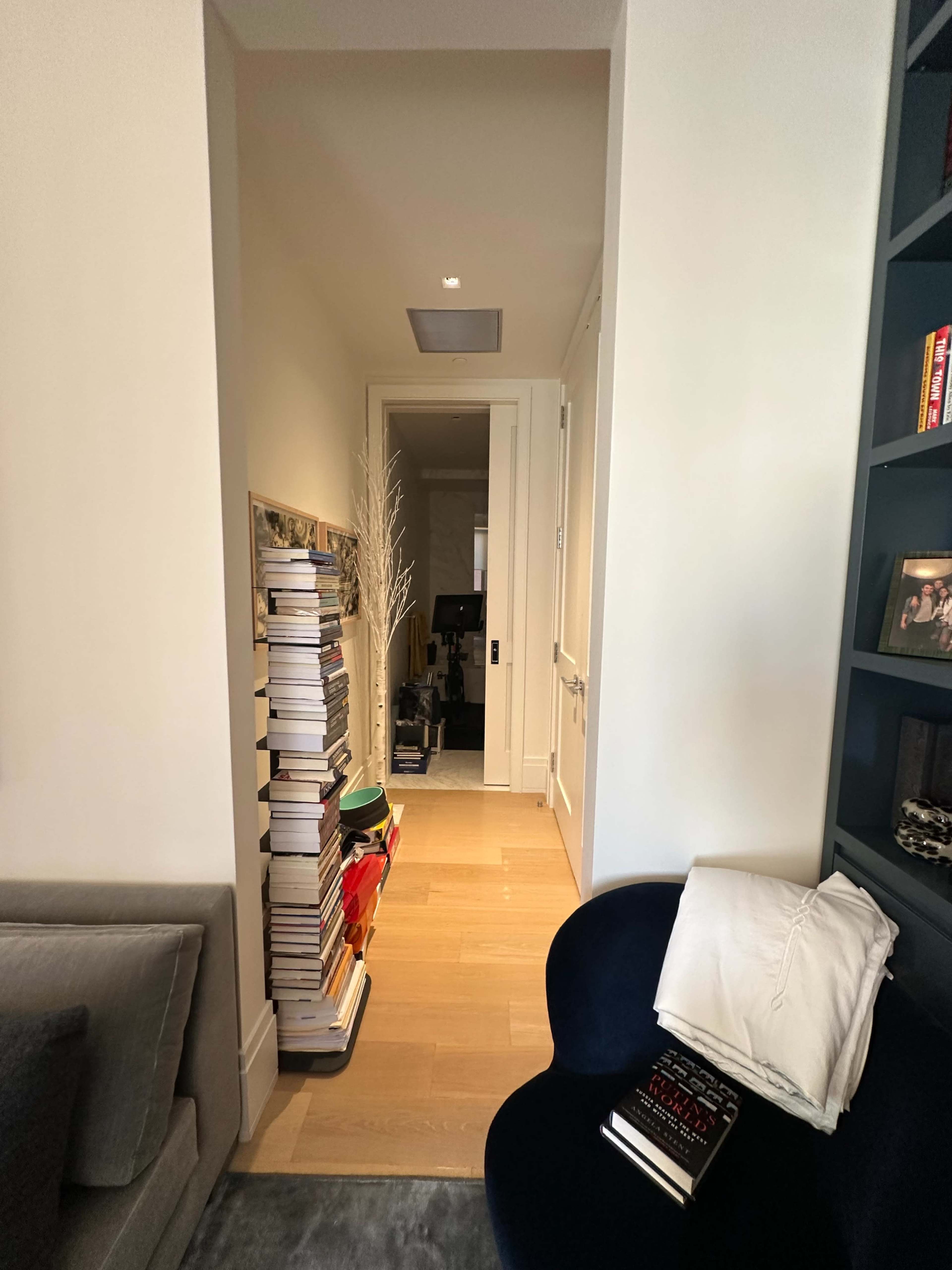 A narrow hallway with a stack of books against one wall leads to a room with a desk and computer at the end.