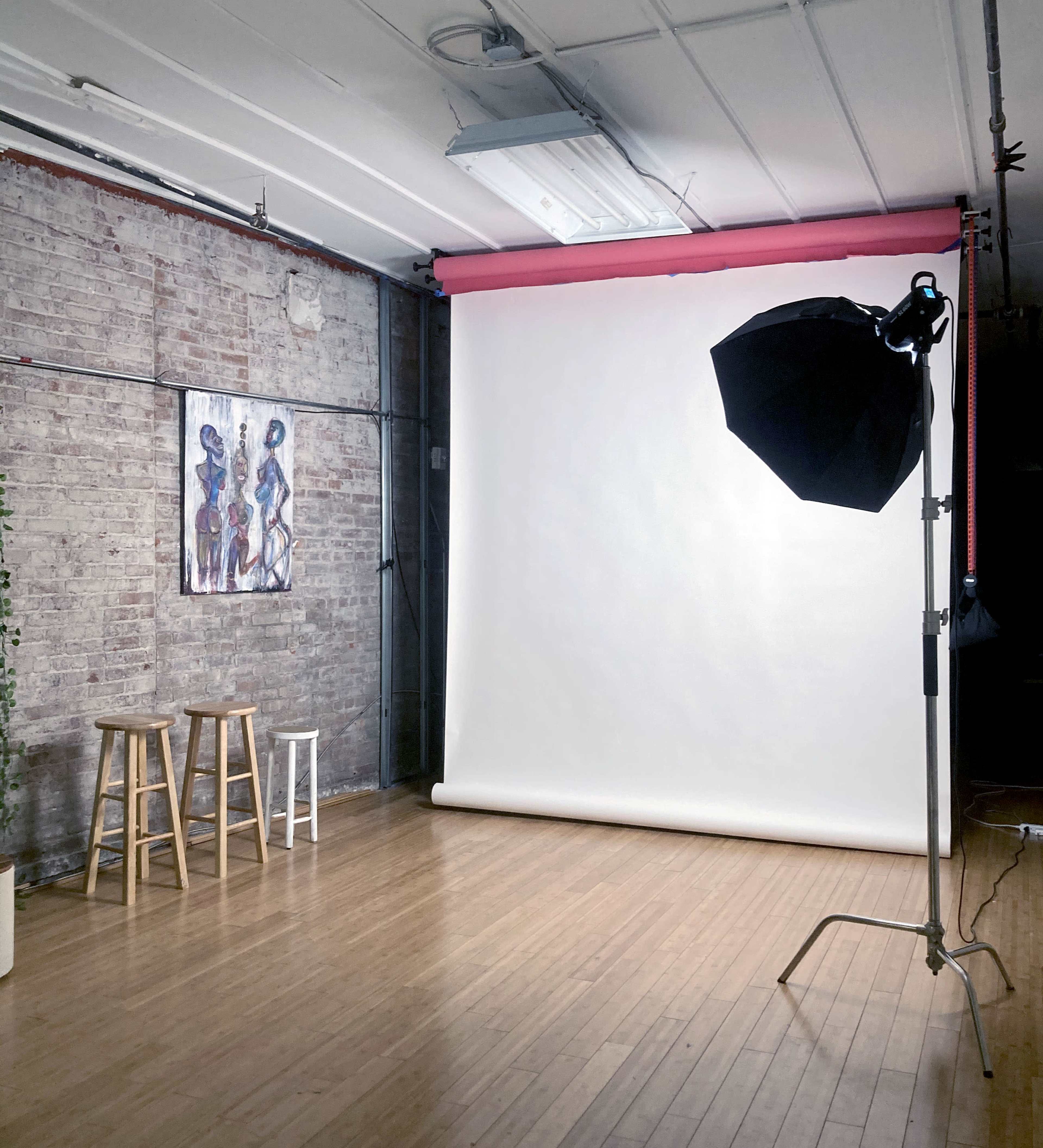 A photography studio with a white backdrop, lighting equipment, and two stools against a brick wall featuring a painting.
