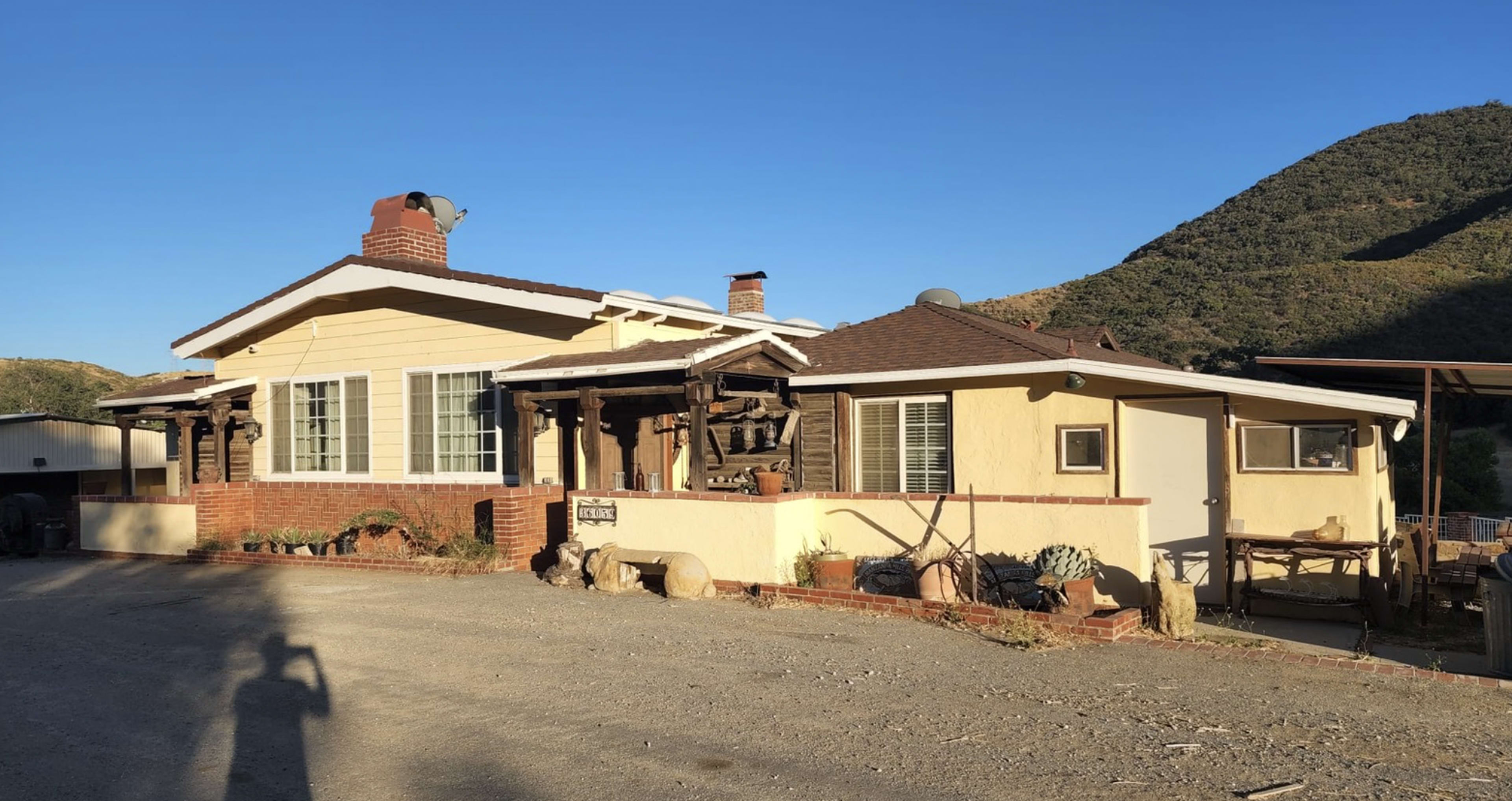 A yellow and brown single-story house with a brick chimney is situated next to a gravel driveway and surrounded by hills.