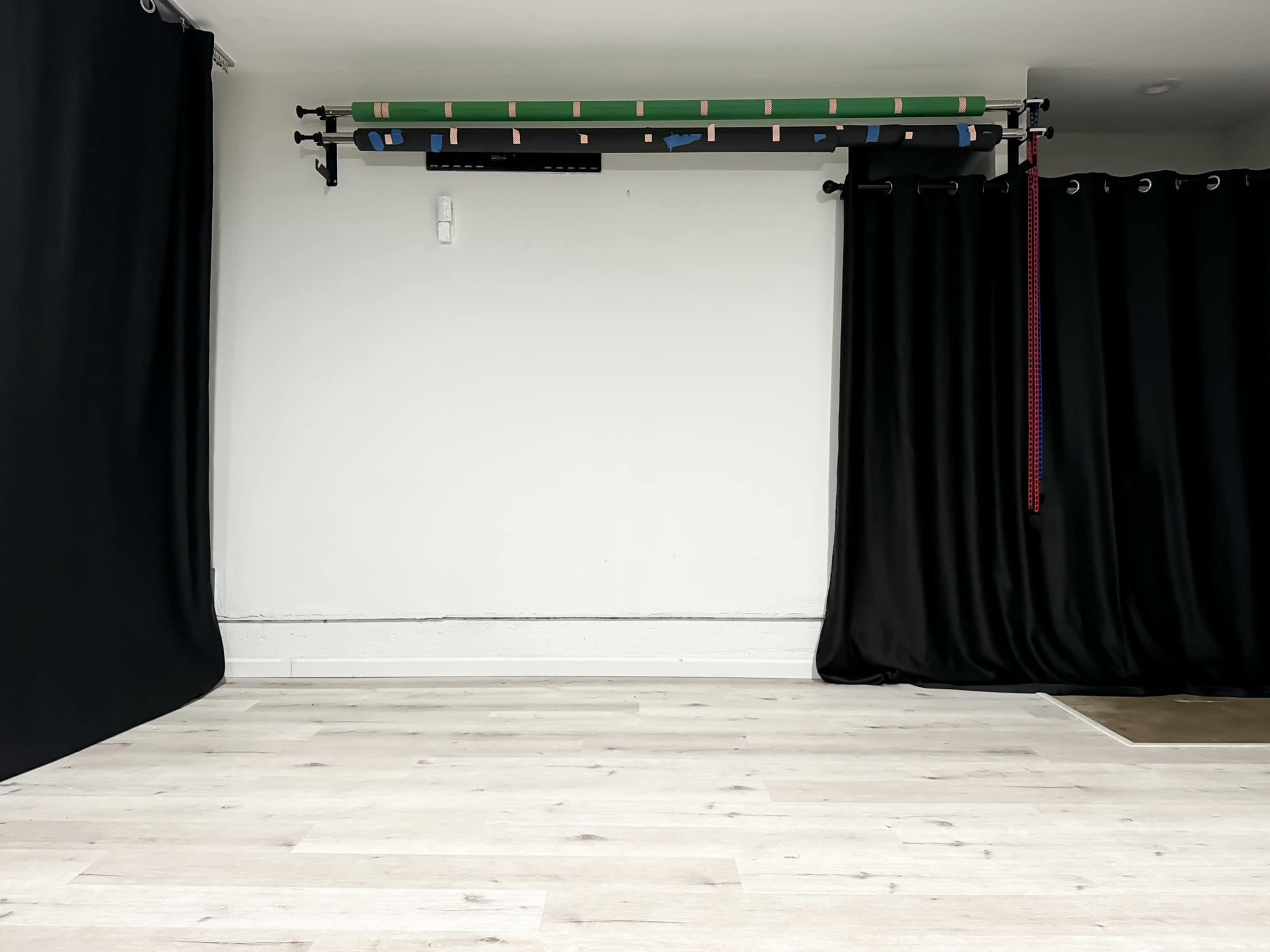 The image shows an empty room with light wooden flooring, a plain white wall, and black curtains on one side, along with equipment mounted on the wall above.