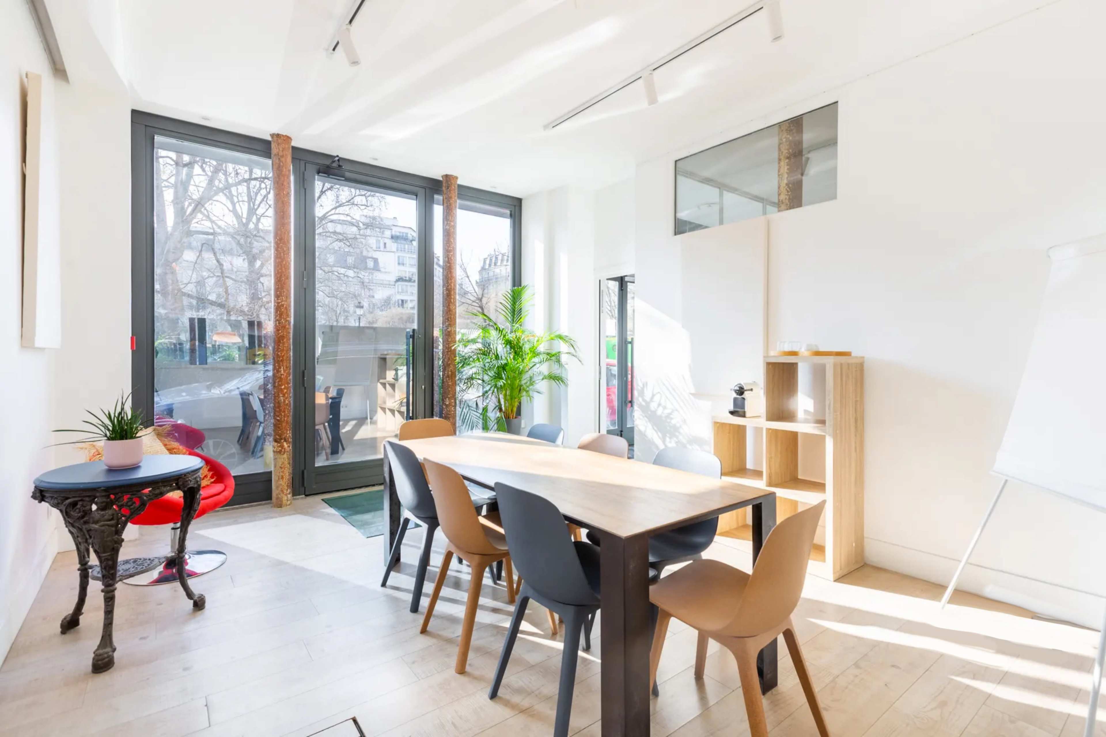 A bright meeting room with a long wooden table surrounded by chairs, large windows overlooking a green space, and a potted plant in the corner.