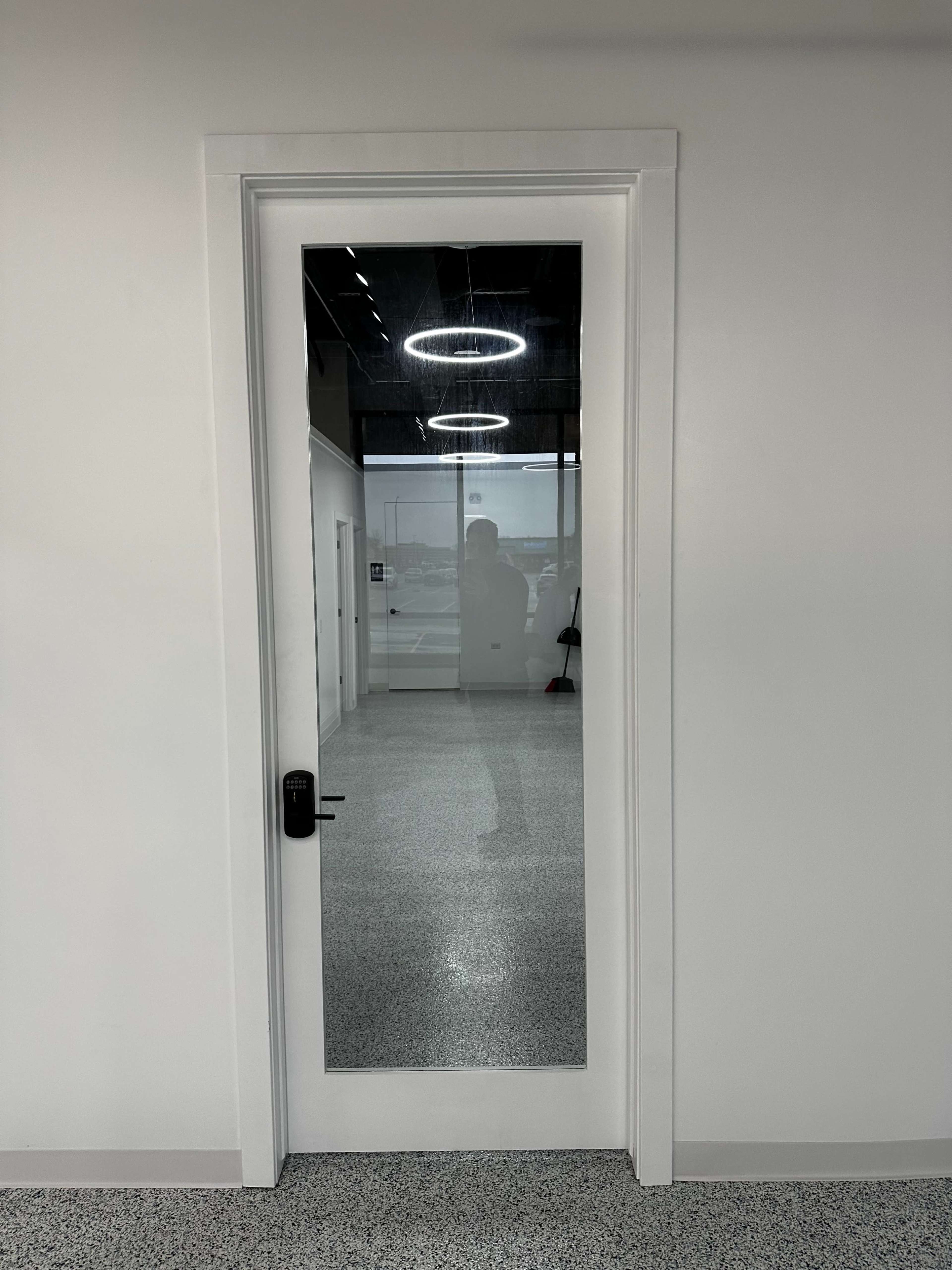 A white door with a glass panel reflects the interior of a room with a polished floor and overhead lights.