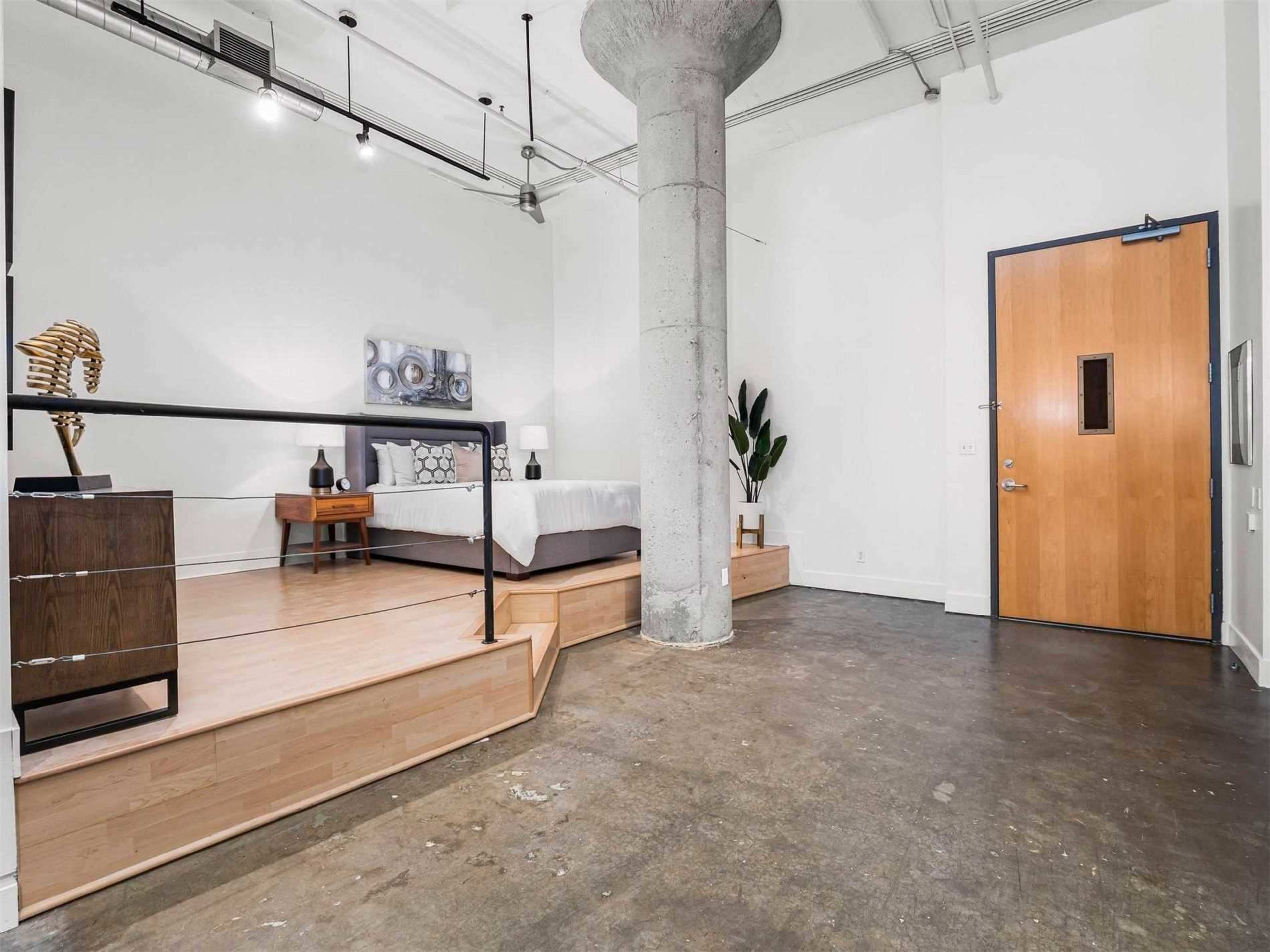 A spacious room features a bed on a raised platform, a concrete pillar, and a door with a glass panel.