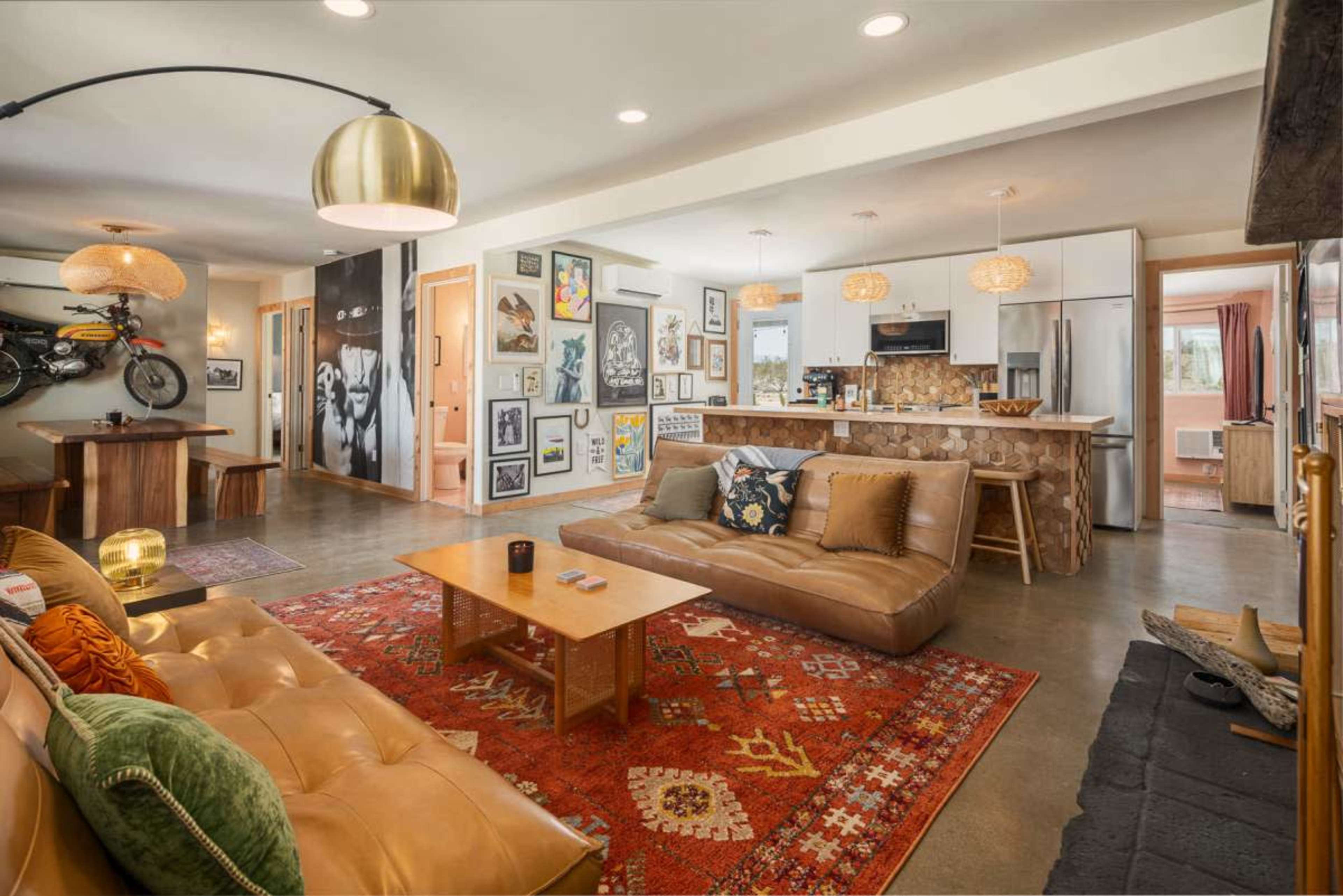 The image shows a modern, open-concept living area featuring a beige leather sofa, a rustic dining table, a kitchen with stainless steel appliances, and walls decorated with various framed artworks.