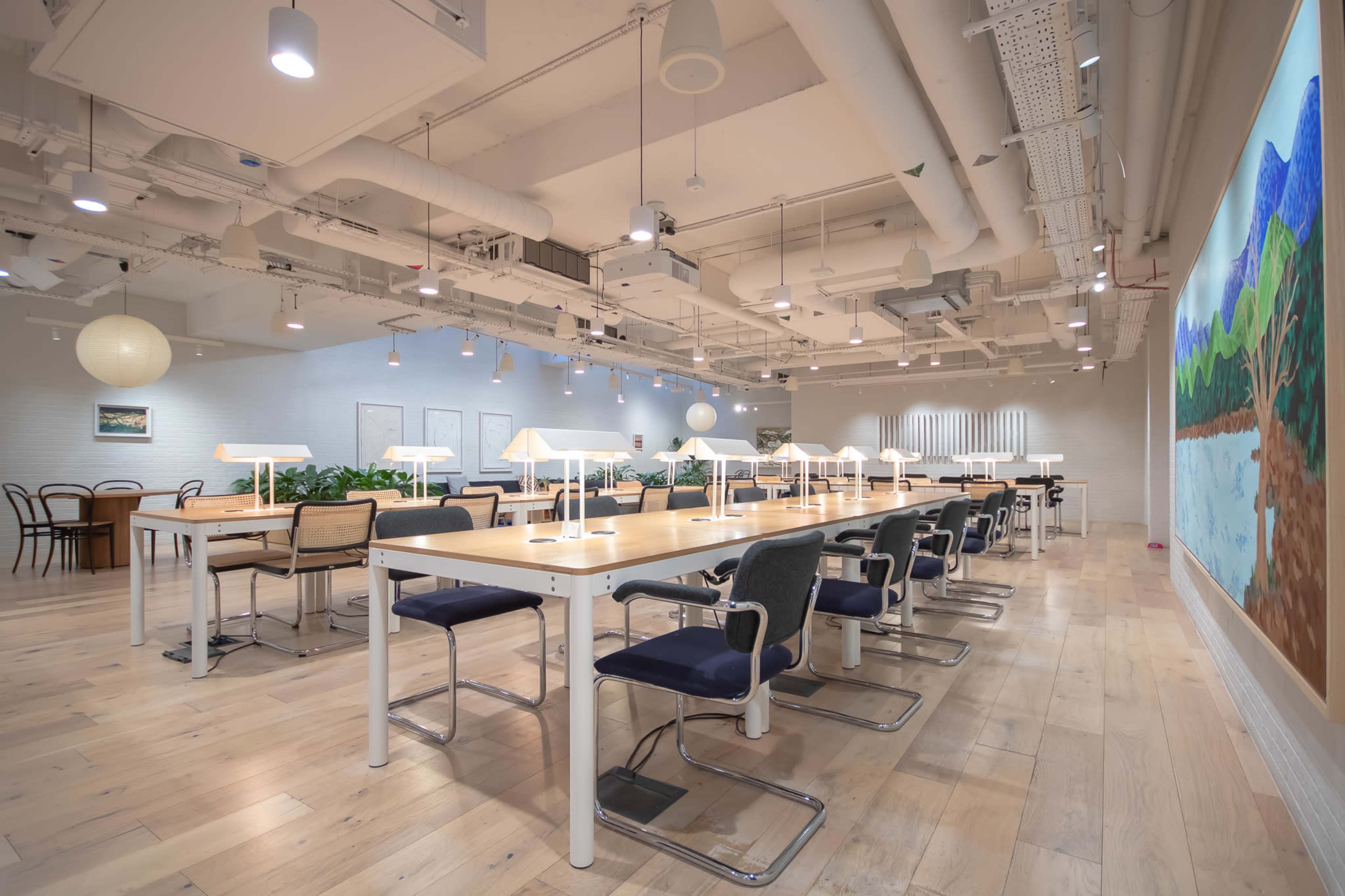 The image shows a spacious, modern office space with several long wooden tables, each equipped with lamps, surrounded by potted plants and white walls.