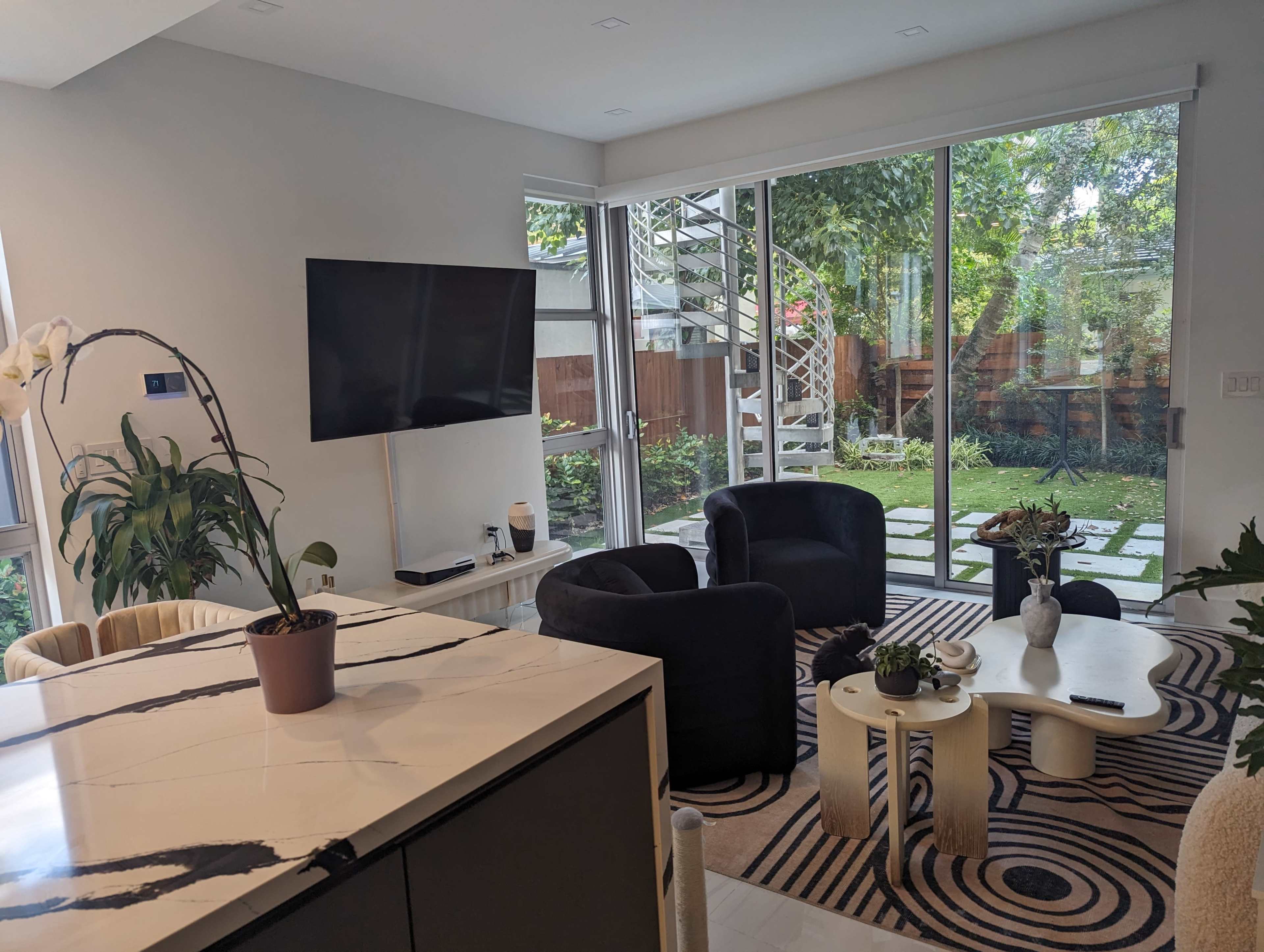 A modern living space with a black television mounted on the wall, two black chairs, and a view of a garden through large glass doors.