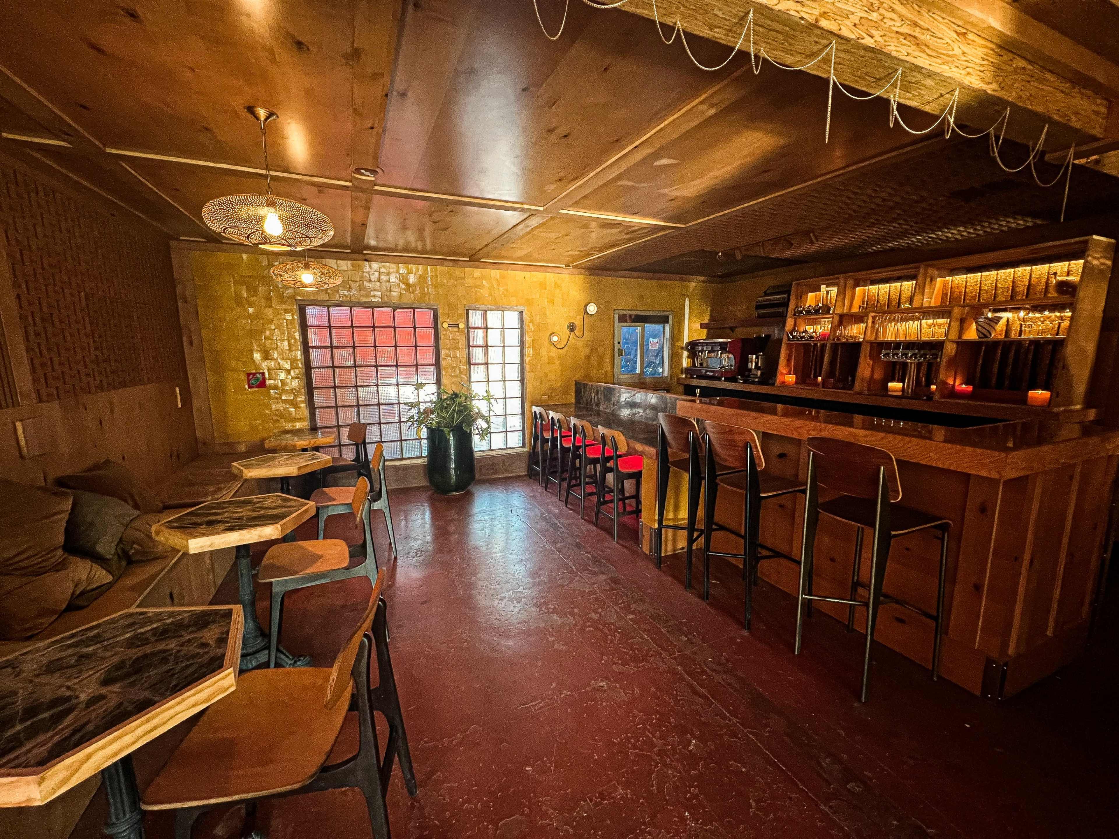 A bar interior with a wooden counter, red accent chairs, and tables, illuminated by warm lighting against a yellow wall.