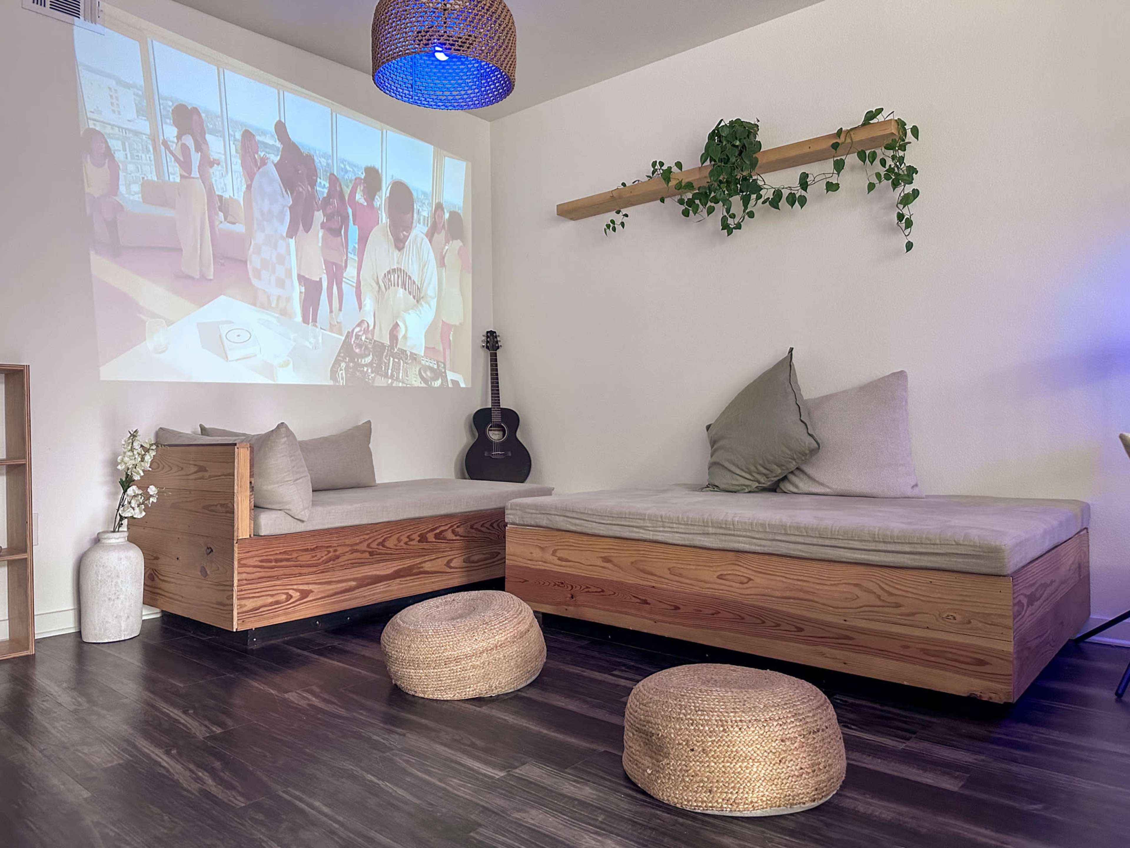 The room features two wooden lounge beds with cushions, woven stools, and a wall-mounted shelf, while a projector displays a video of people in a bright, airy space.