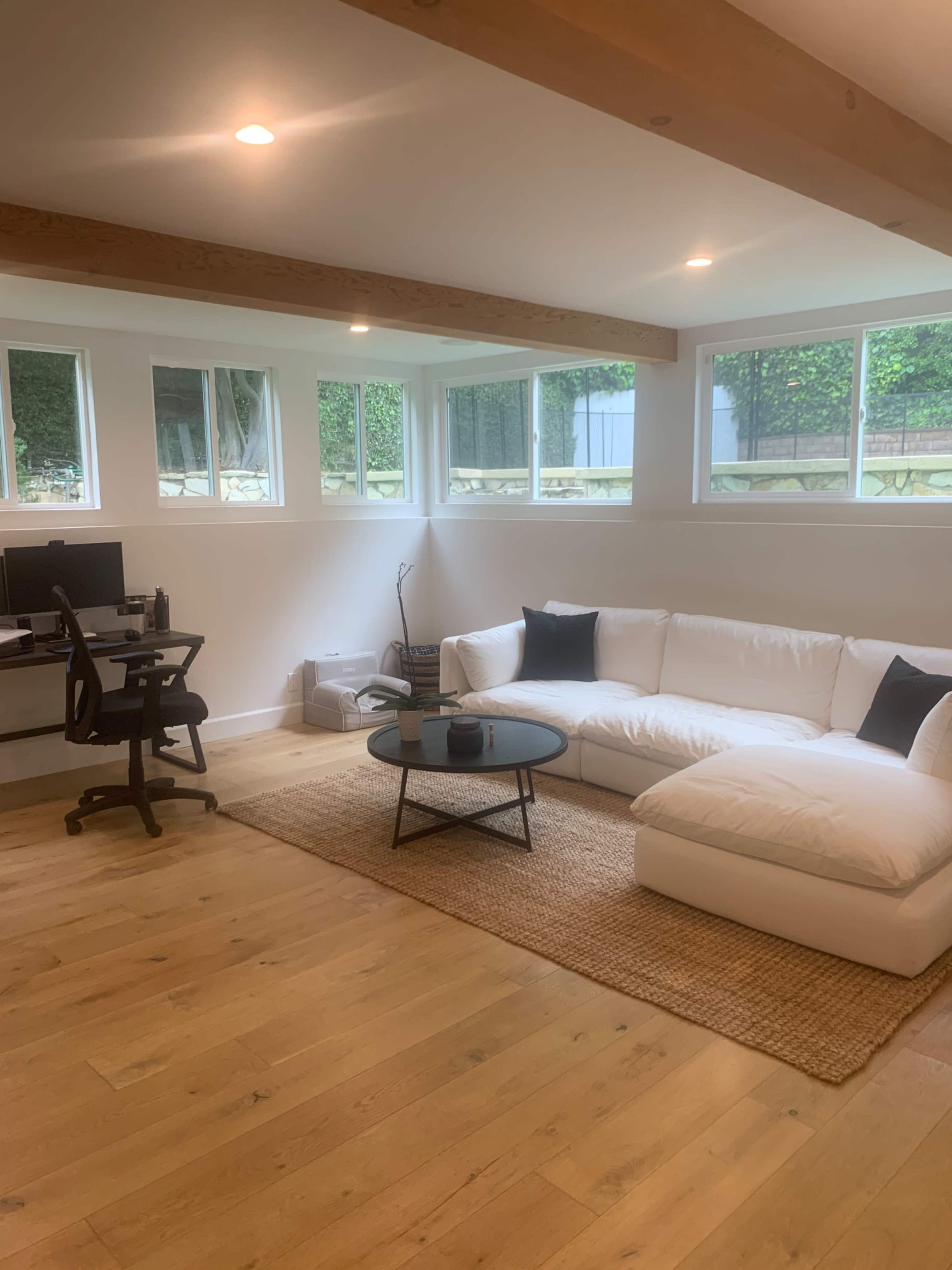 A bright, minimalist room features a white sectional sofa, a round black coffee table, and a desk with two monitors near large windows.