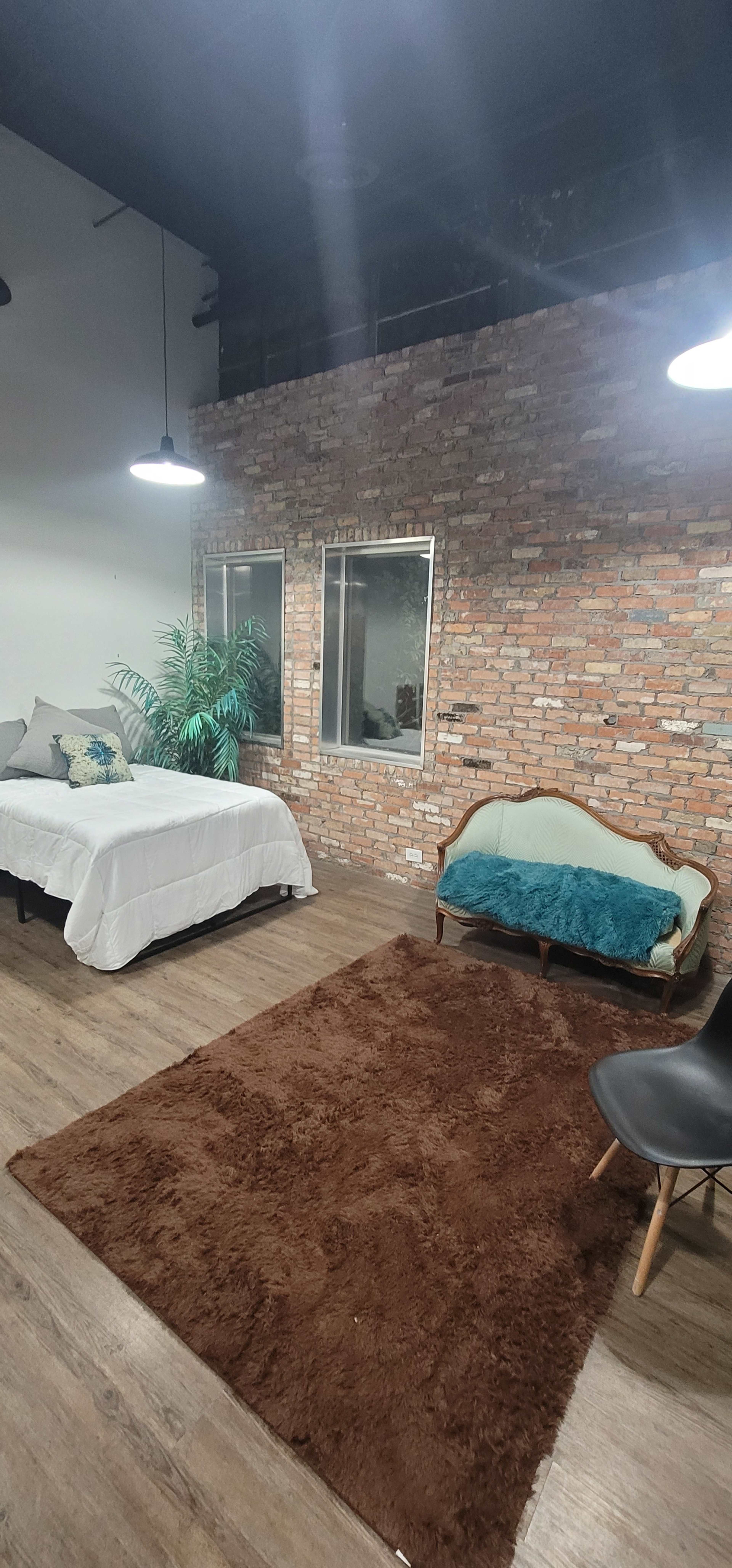 A room with a bed, a vintage-style sofa, a plant, and large windows, all set against a brick wall and wooden flooring.