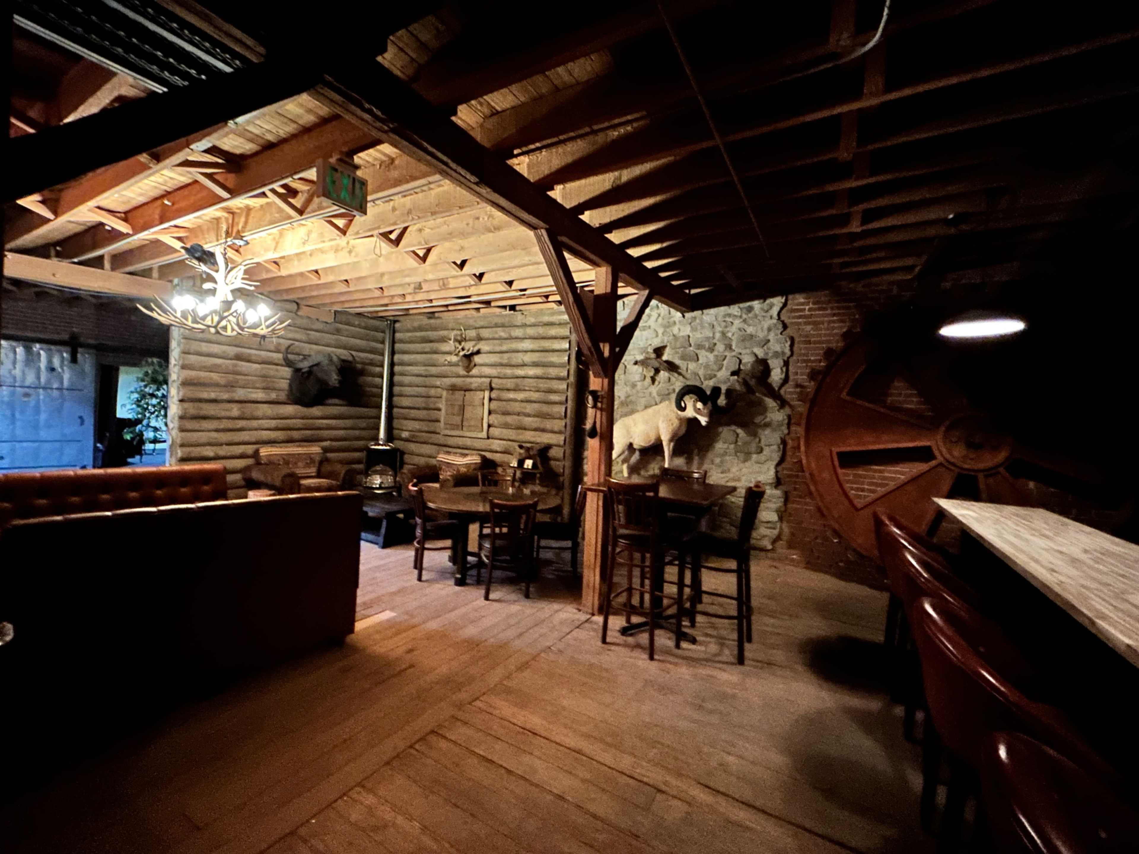 The image shows a rustic interior of a wooden lodge with a bar area, tables, and mounted animal heads on the walls.