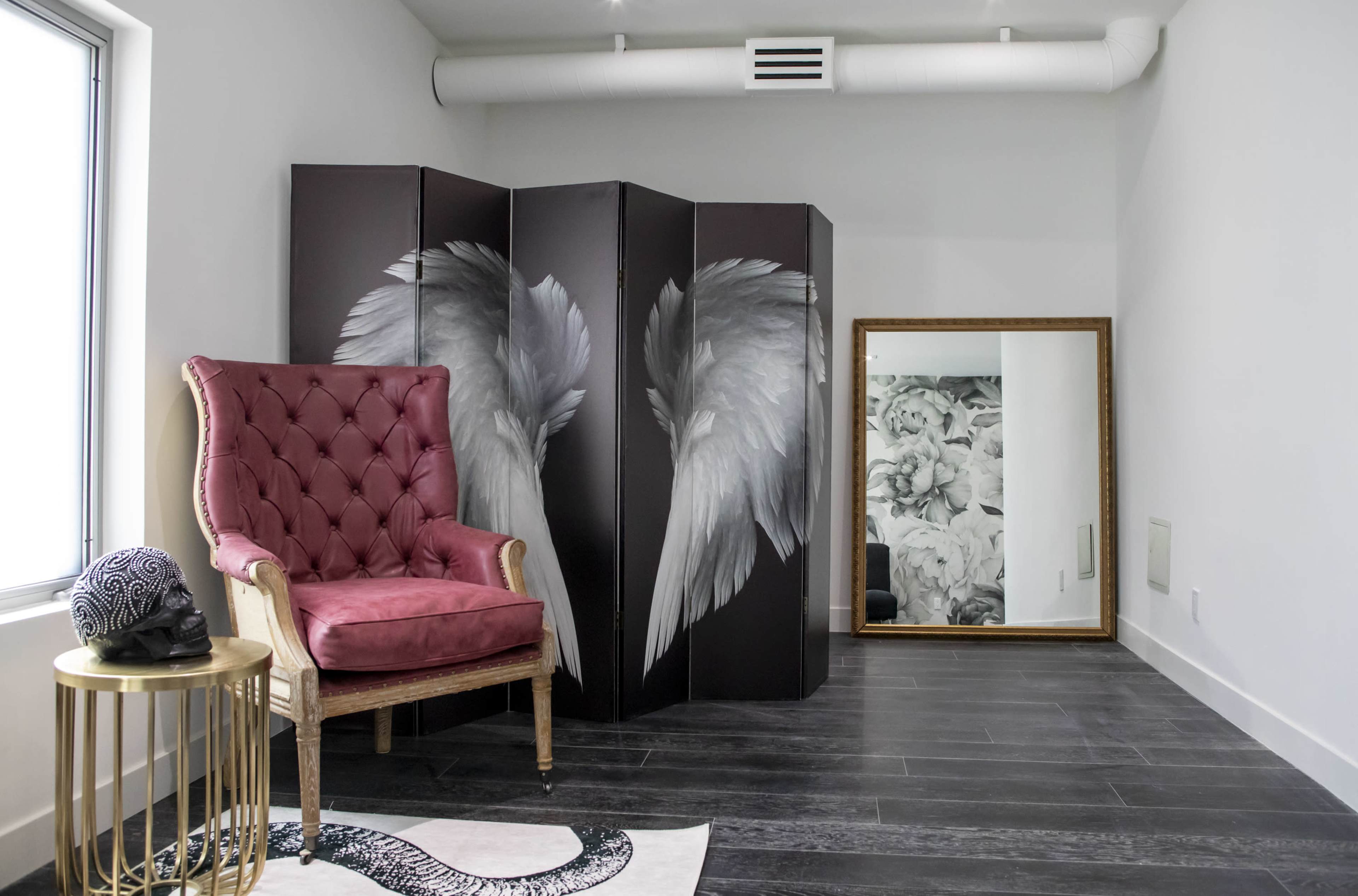The room features a pink armchair next to a gold side table, with a folding screen displaying wing artwork and a large ornate mirror reflecting a decorated wall.