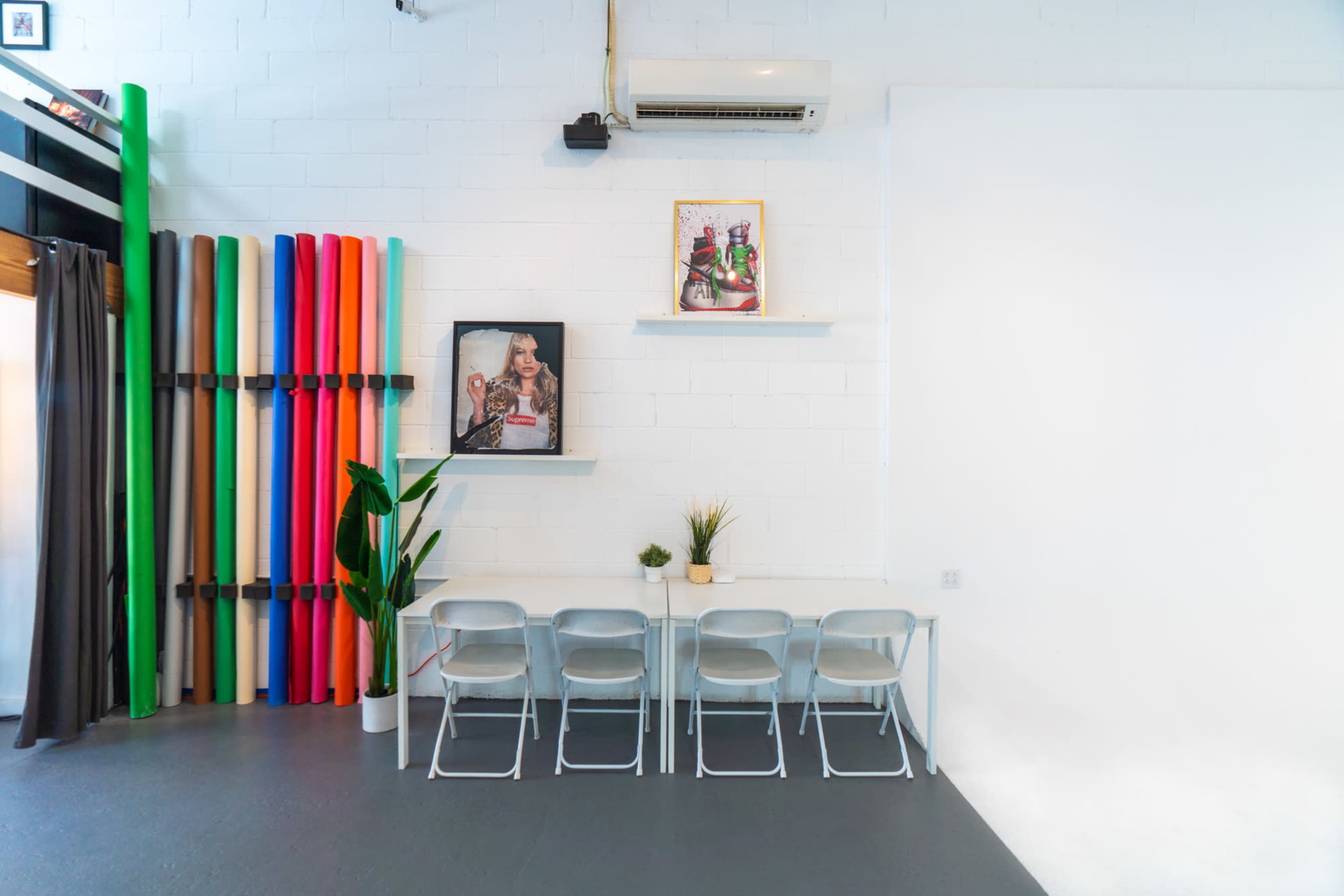 The image shows a bright workspace with a long table and six chairs, colorful rolls of material on the wall, and decor including framed images and potted plants.