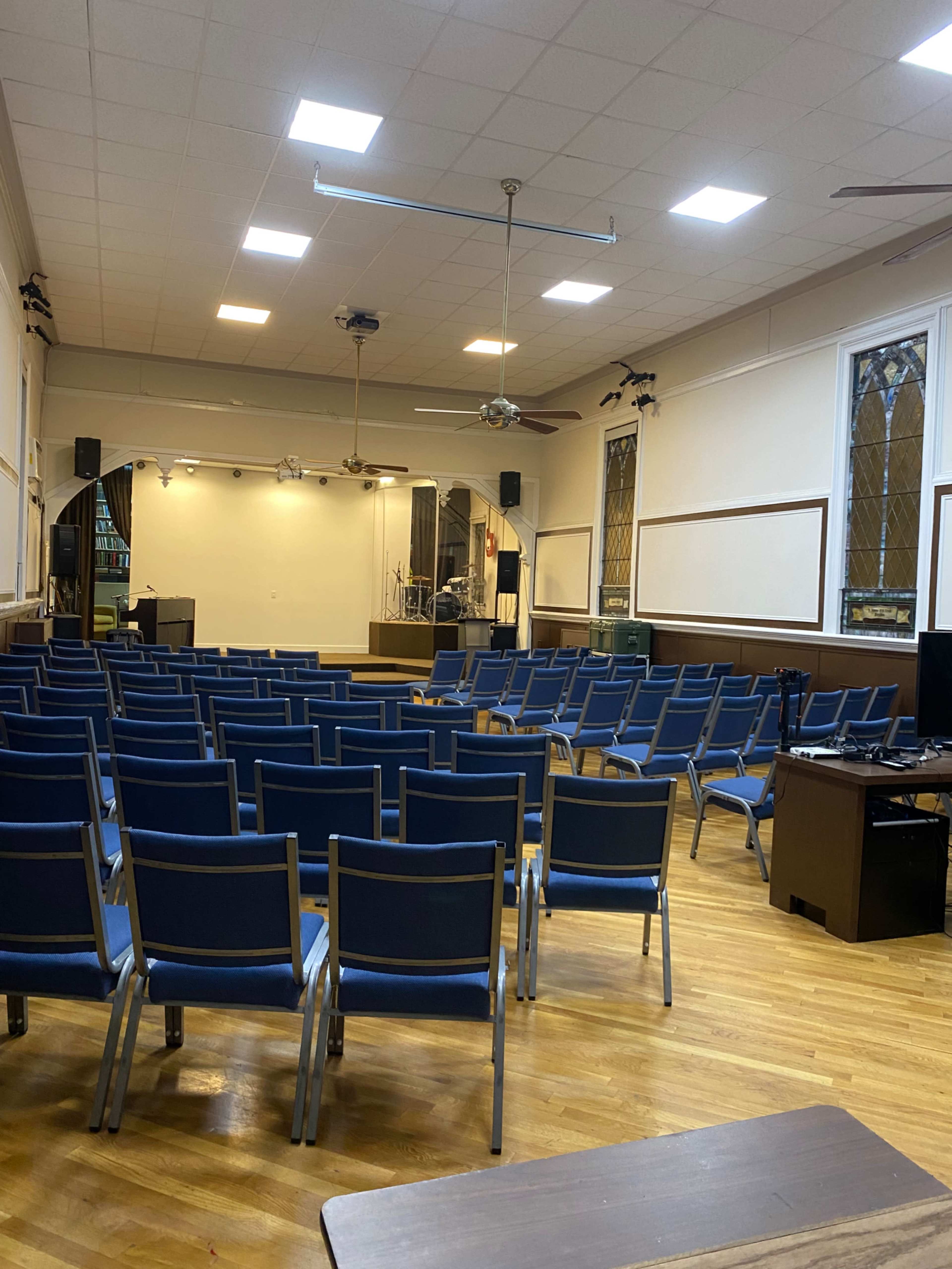 A mostly empty auditorium with rows of blue chairs facing a stage and illuminated by natural light from large windows.
