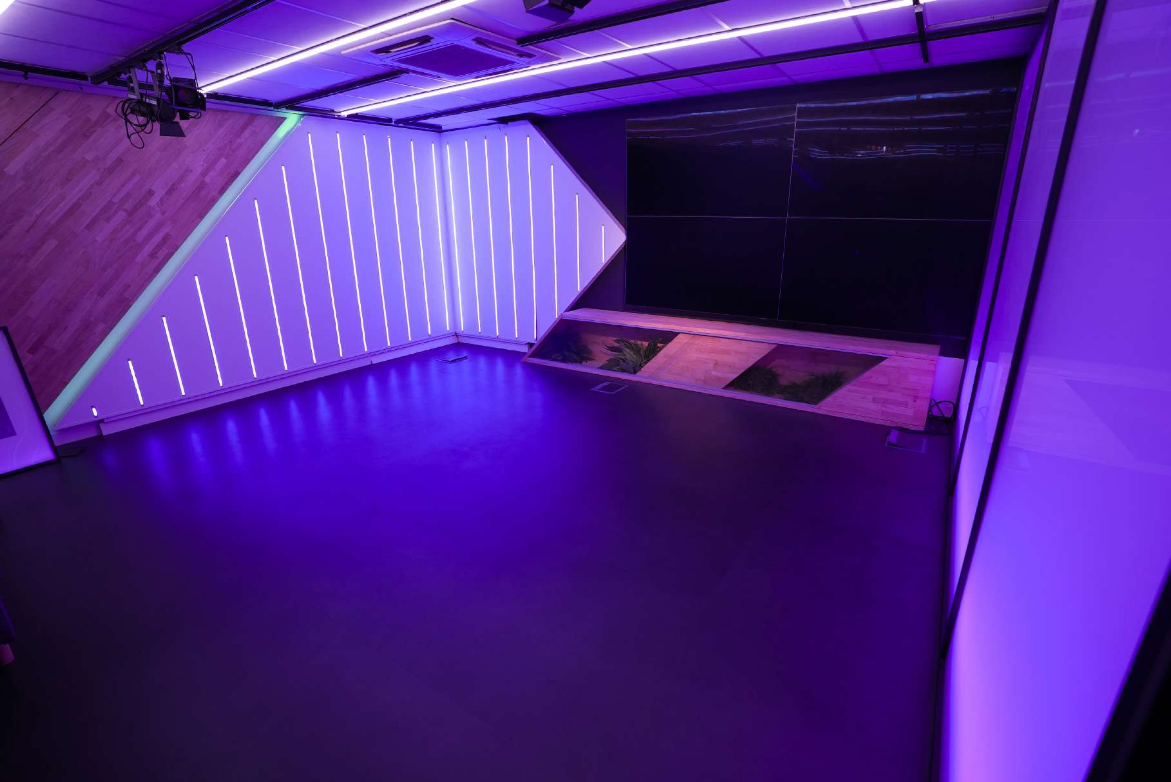 The image shows a modern studio space with angular walls illuminated by purple LED lights and a large digital screen on one side.