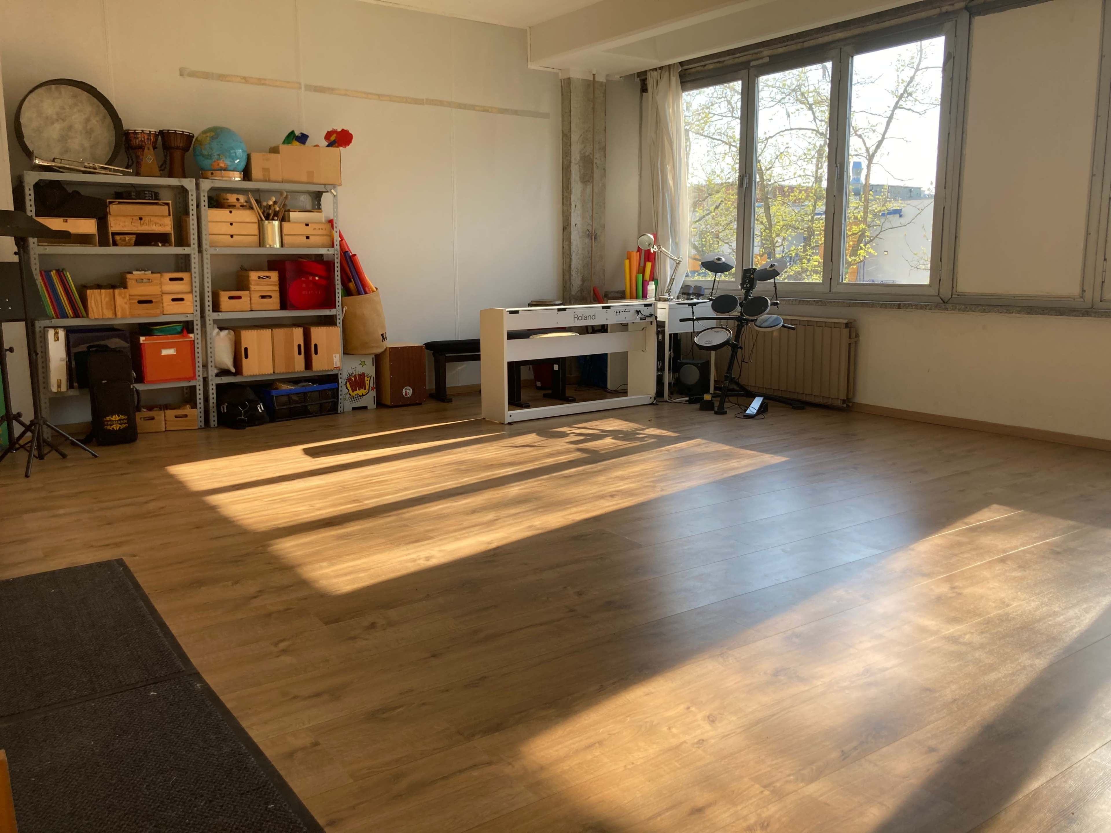 A spacious room with wooden floors, bright sunlight streaming in through large windows, and various storage boxes and musical instruments against the walls.